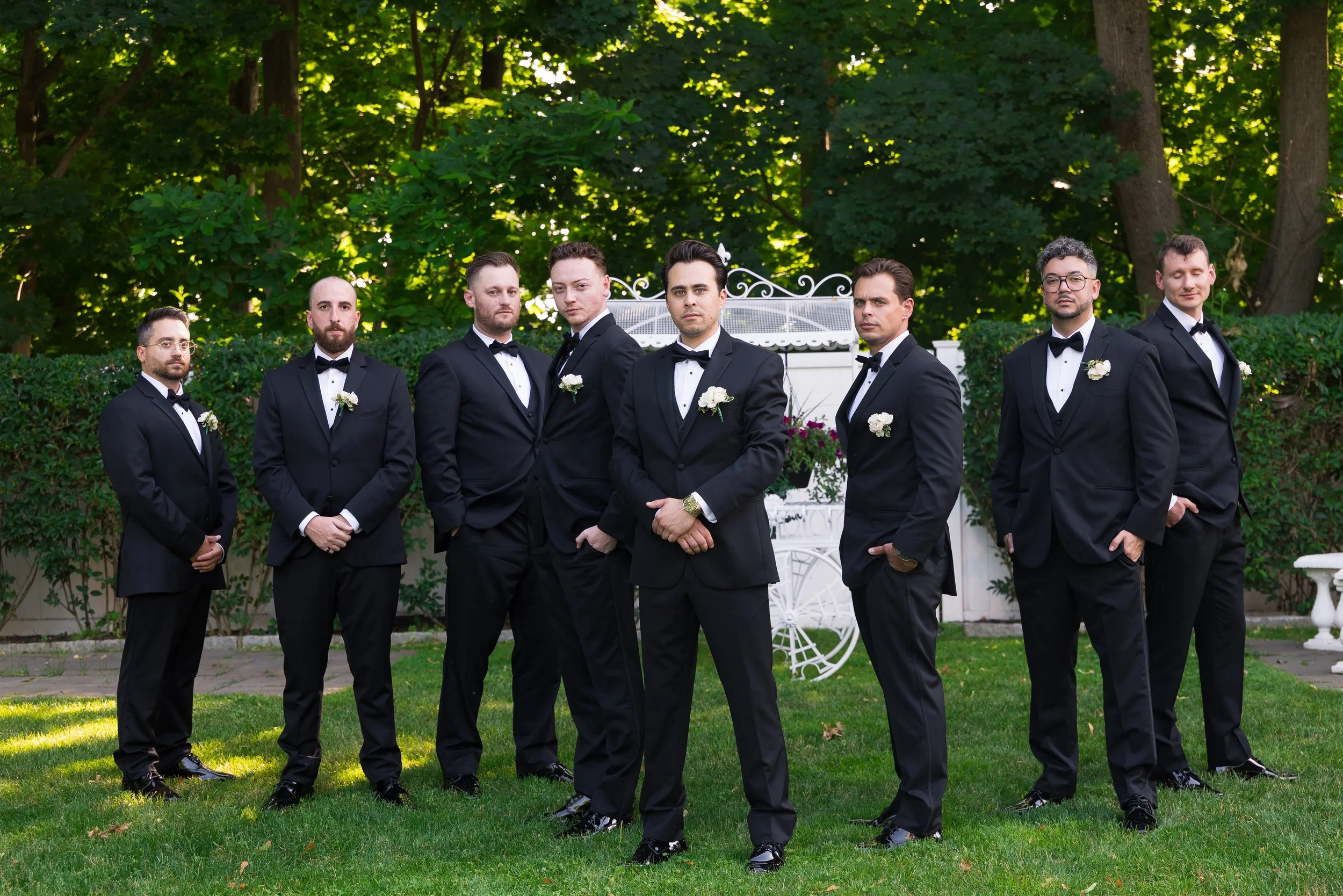 A group of eight men dressed in black tuxedos with white shirts and black bow ties, standing outside on a lawn with trees and greenery behind them. They appear to be at a formal event or wedding.