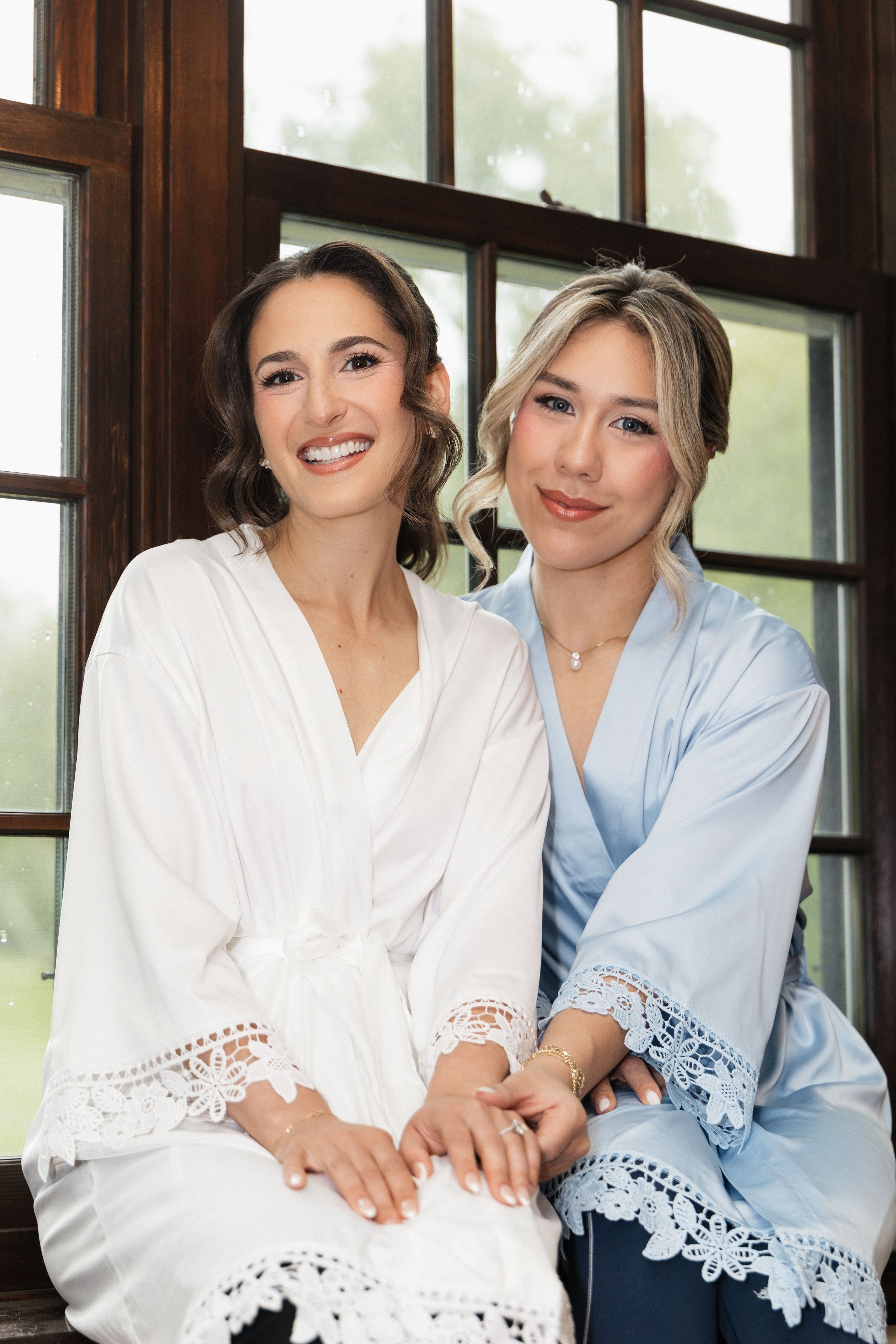 Two women sitting close together in front of large window, smiling at the camera. One woman has dark hair and is wearing a white dress with lace details, and the other has blonde hair and is wearing a light blue dress with lace details.