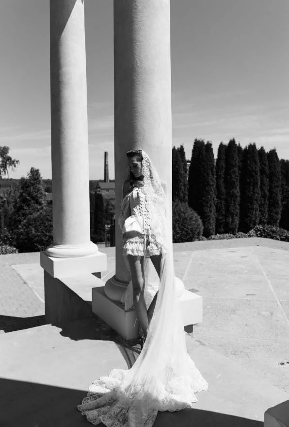 A woman in vintage lace lingerie and a long veil, standing between two large classical columns outdoors on a sunny day, with trees and a distant factory in the background.