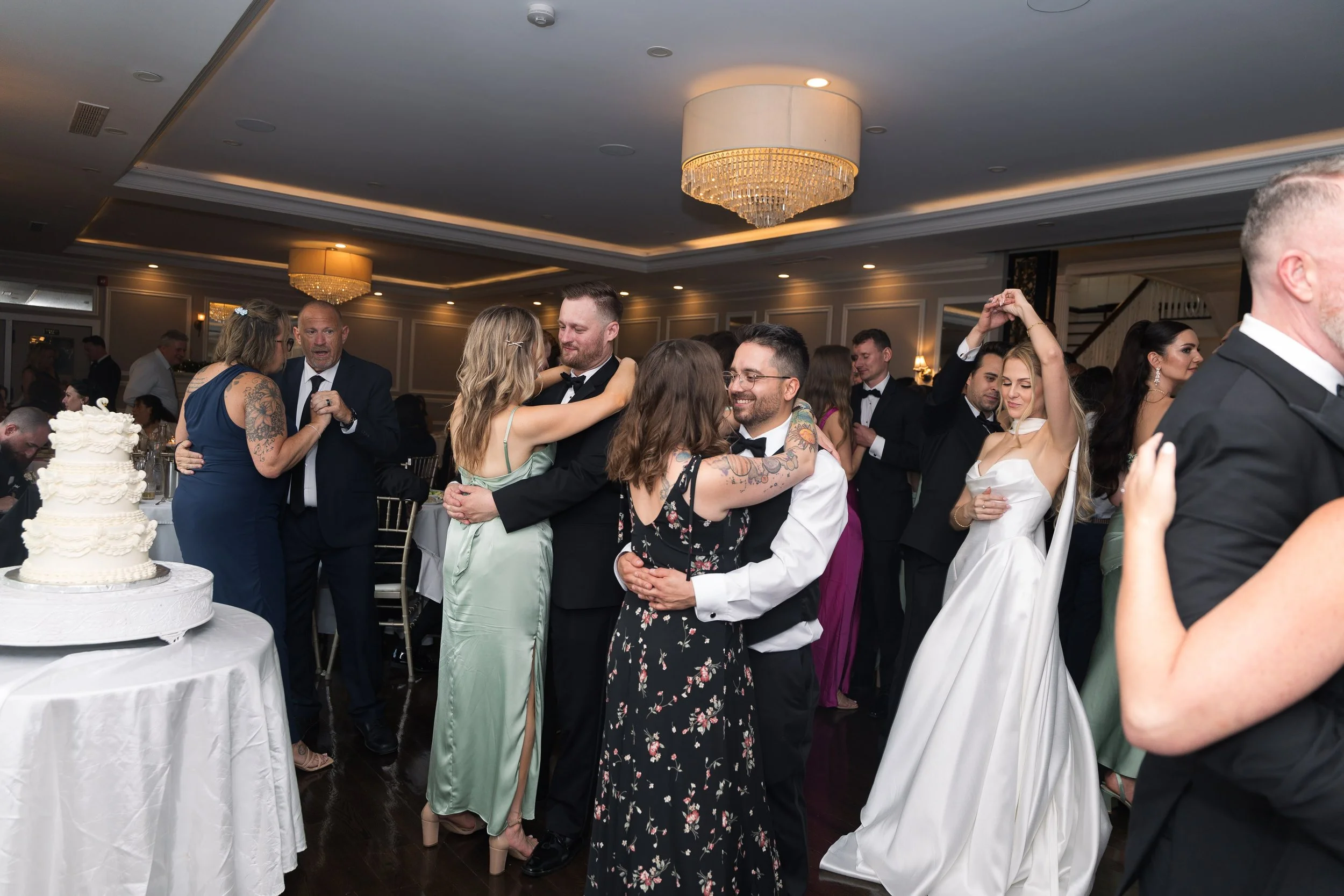 People dancing and celebrating at a formal event, with a wedding cake visible on a table in the foreground.