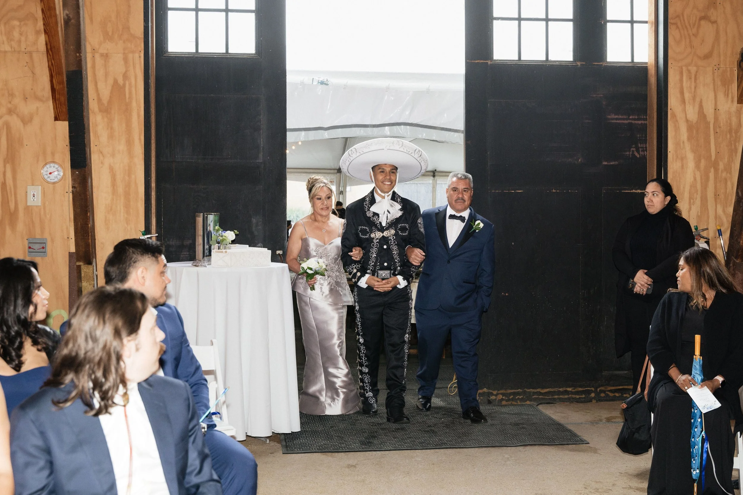 A wedding ceremony with a man and woman walking down the aisle, with the man dressed in traditional Mexican Mariachi attire and a sombrero, and the woman in a wedding dress. Guests seated on either side watch them as they walk.