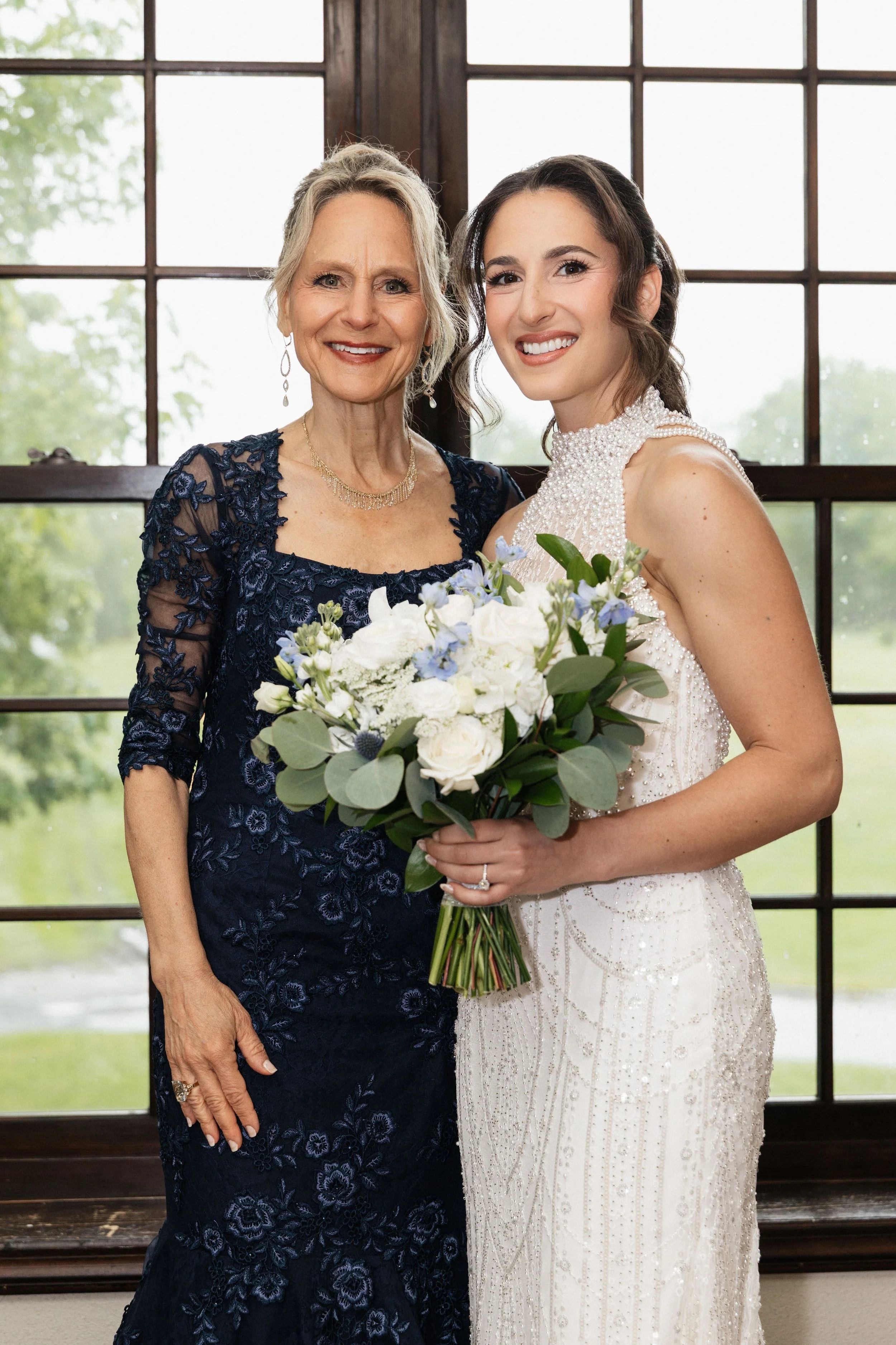 A woman in a navy lace dress stands next to a woman in a white wedding gown, holding a bouquet, inside a room with a large window showing greenery outside.