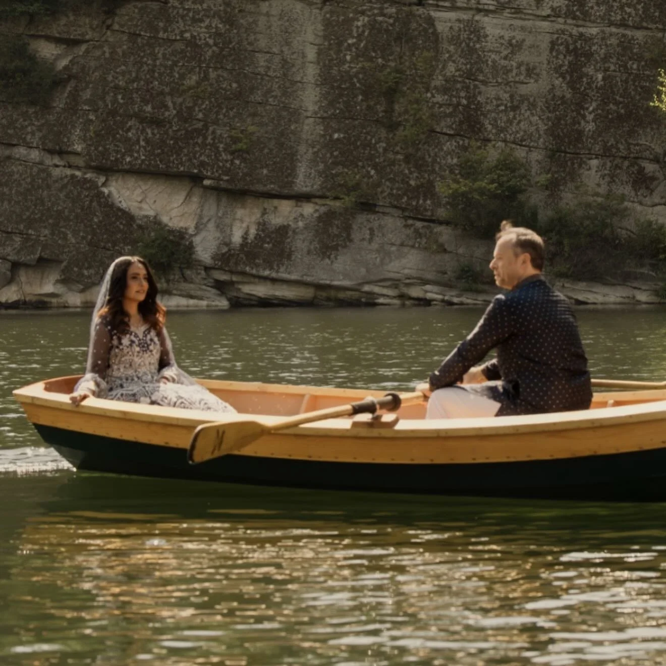 A couple in a small boat on a river surrounded by rocky cliffs, with the woman wearing a veil and dress and the man in a dark shirt, engaging in a romantic moment.