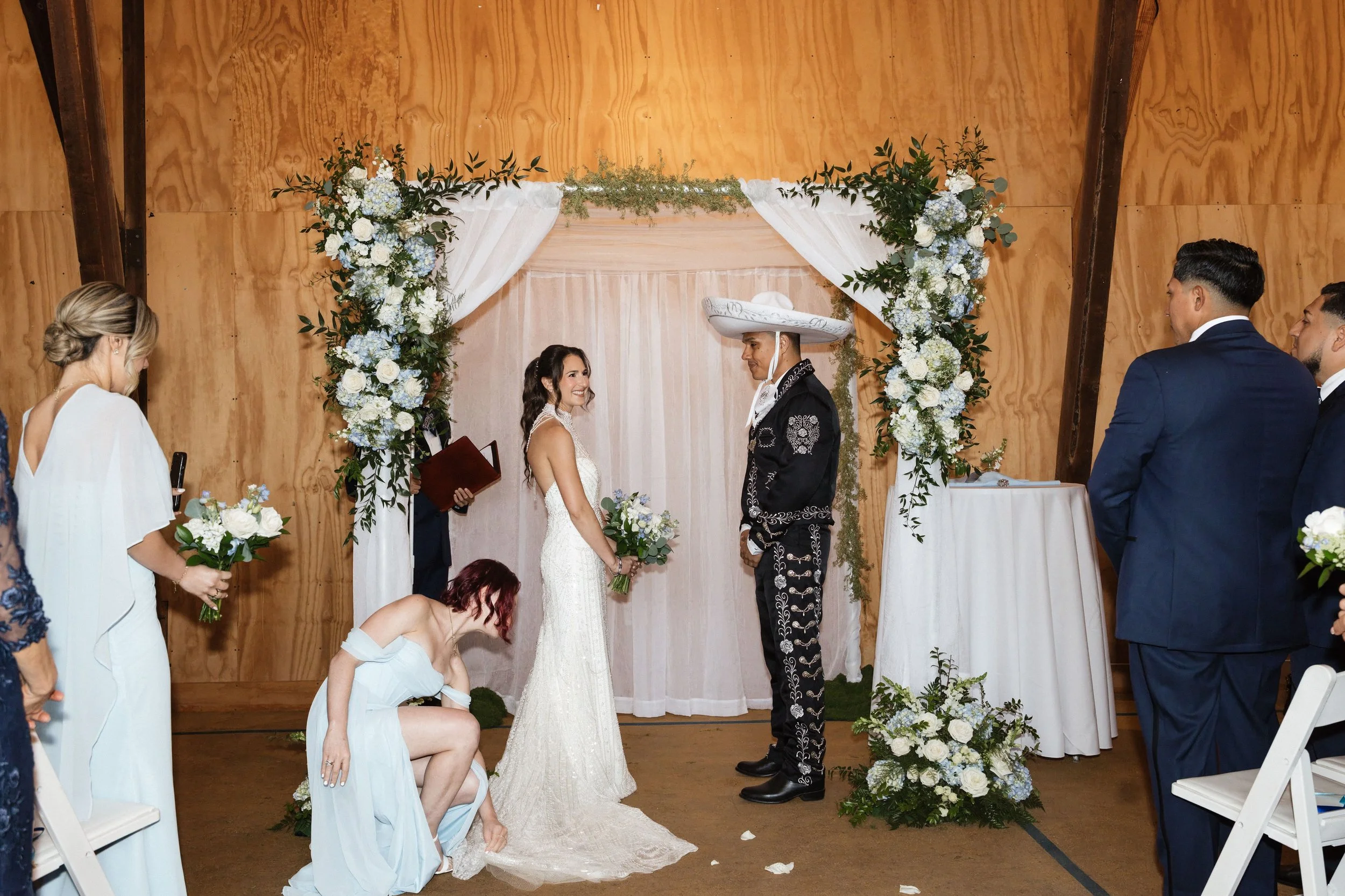 A wedding ceremony with a couple exchanging vows under a floral arch with white and blue flowers, surrounded by guests, inside a wooden venue.
