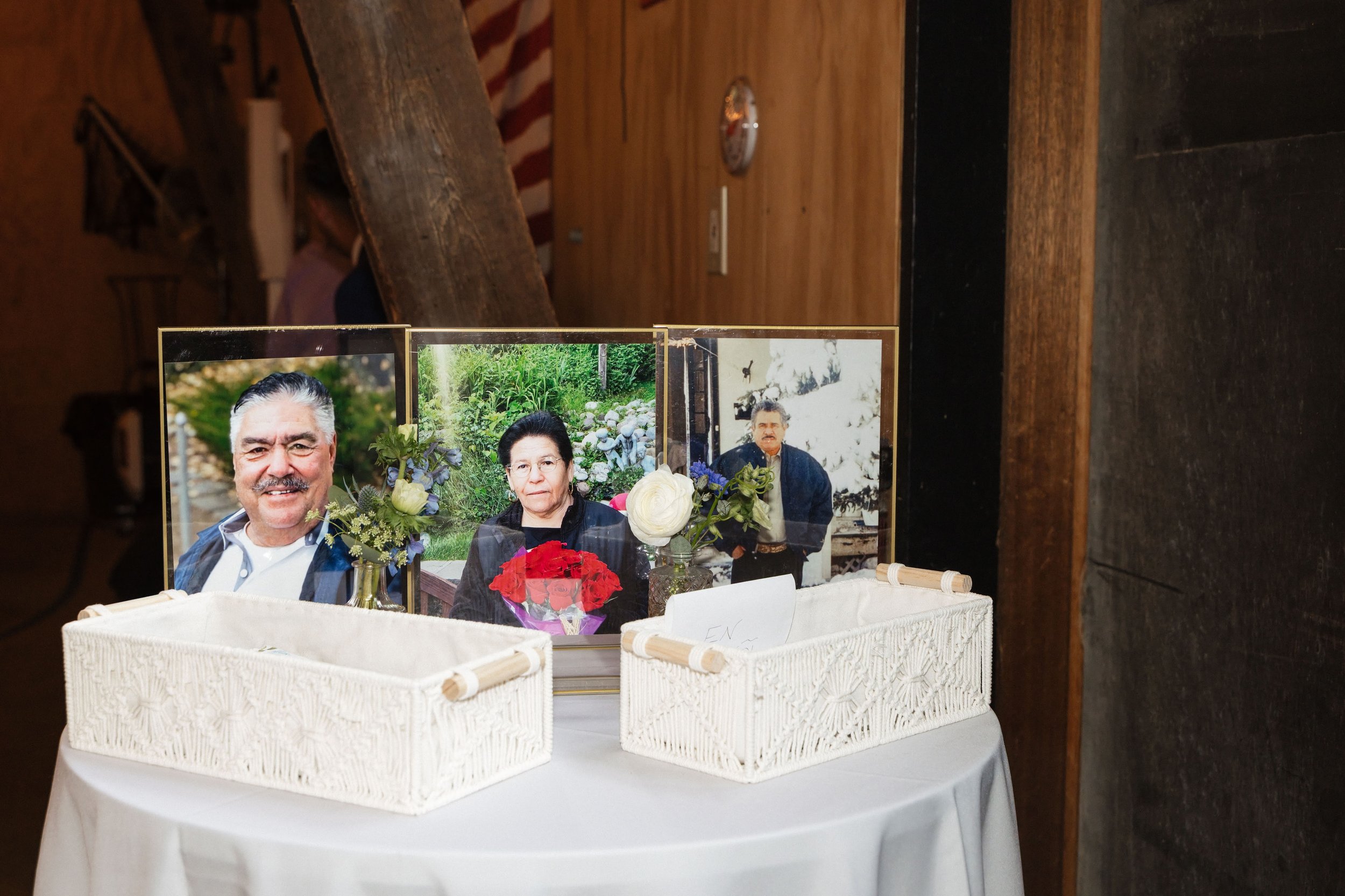 A display with framed photographs of an elderly man and woman, and a man, accompanied by flowers in vases, in a white wicker basket on a table covered with a white cloth.