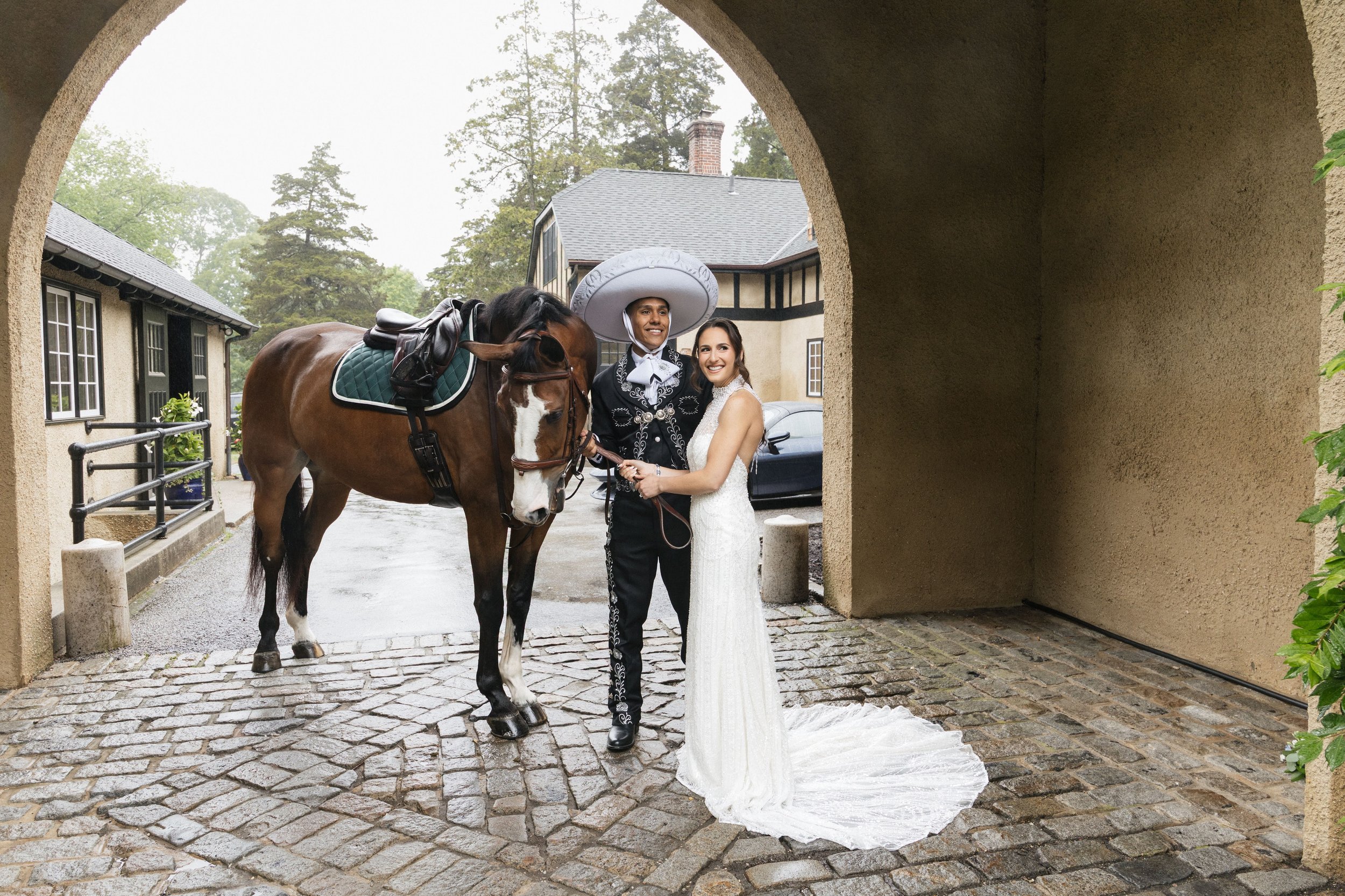 A bride and a man dressed as a mariachi, holding a horse's reins, standing at a wedding ceremony location under an archway with buildings and trees in the background.