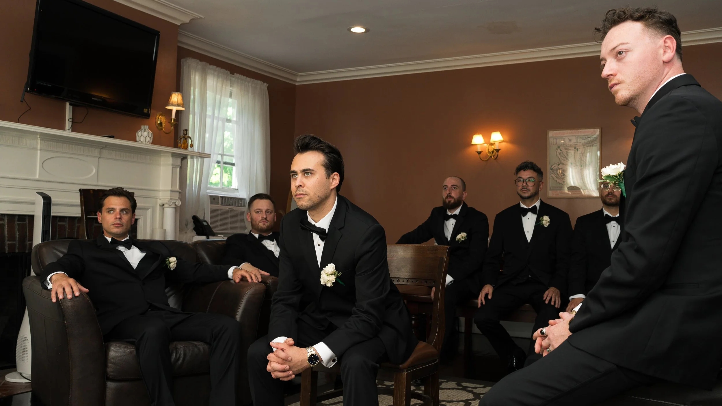 A group of men dressed in formal tuxedos, with some seated on a sofa and others standing or sitting at a table in a living room, during a pre-wedding gathering or ceremony.
