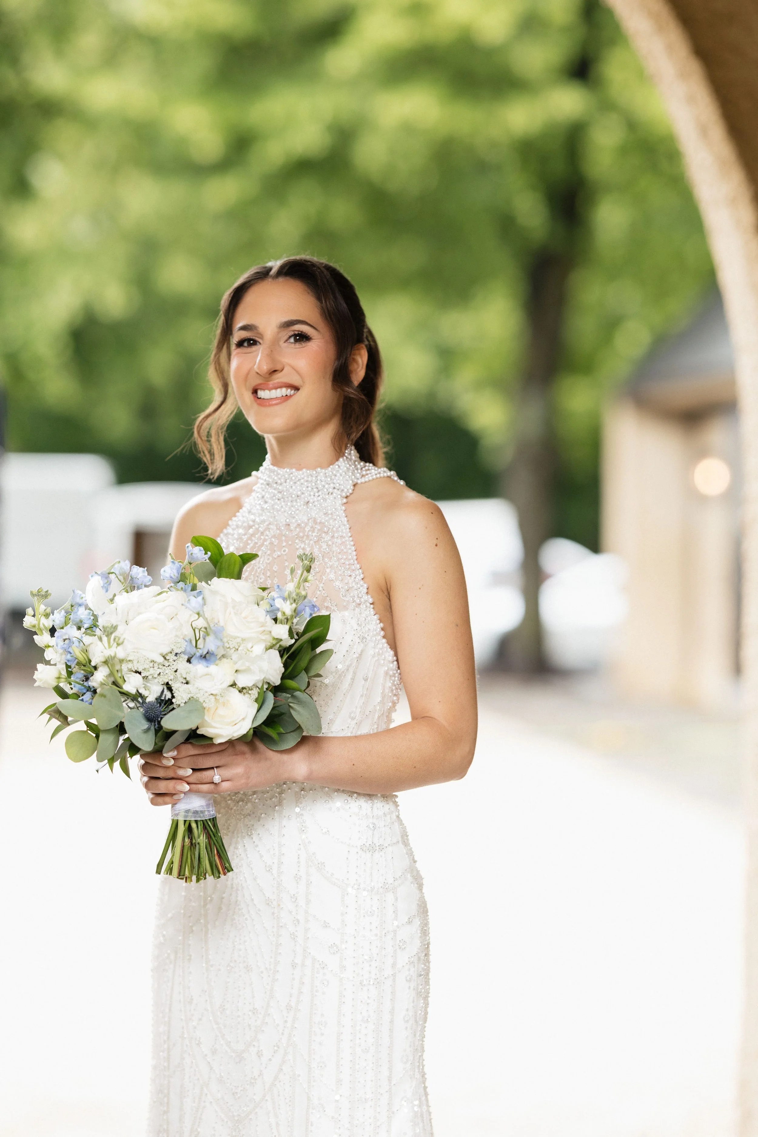A bride in a white wedding dress holding a bouquet of white and light purple flowers, outdoors with blurred trees in the background.
