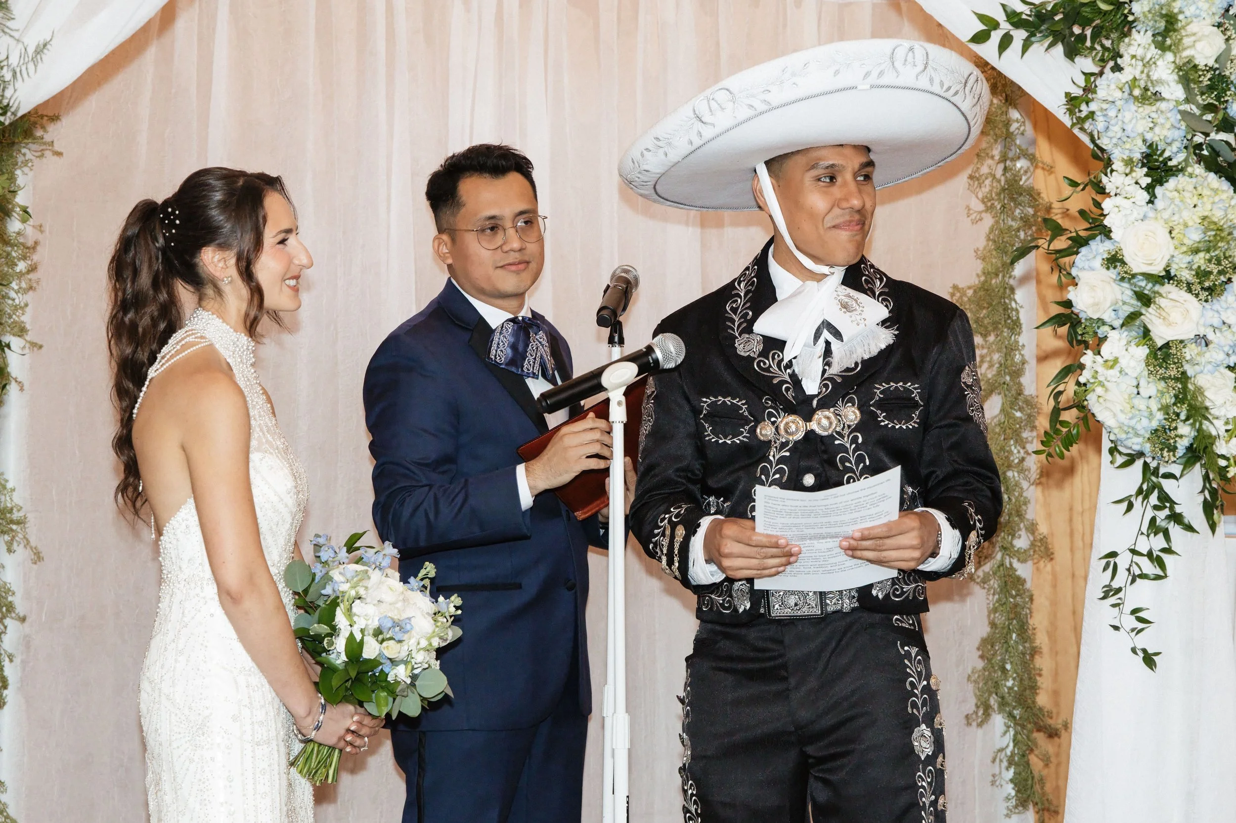 A wedding ceremony with a bride, groom, and officiant dressed in traditional Mexican attire, standing under a floral archway.
