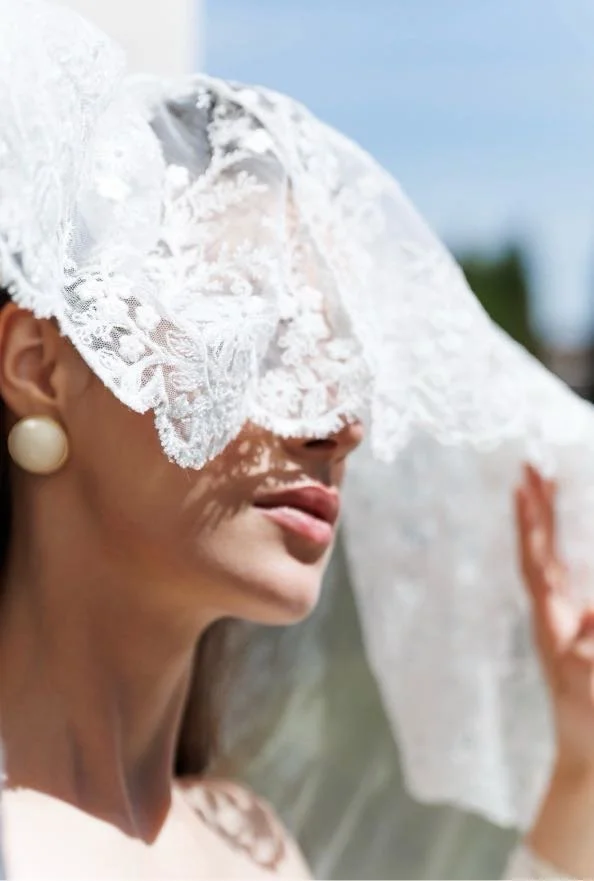 Woman with pearl earrings holding a lace veil over her face outdoors on a sunny day.