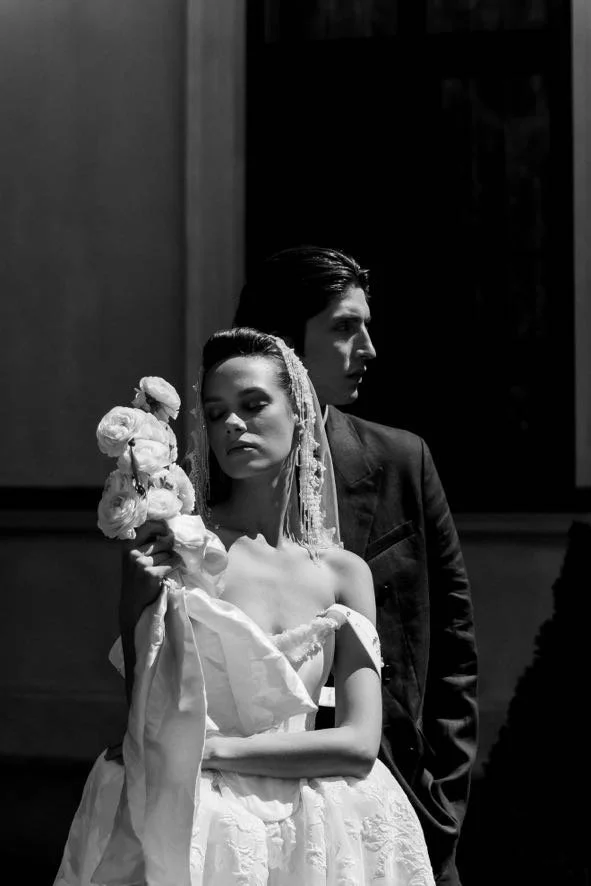 A black and white photo of a bride and groom, with the bride holding a bouquet of flowers and the groom standing behind her, both looking away.