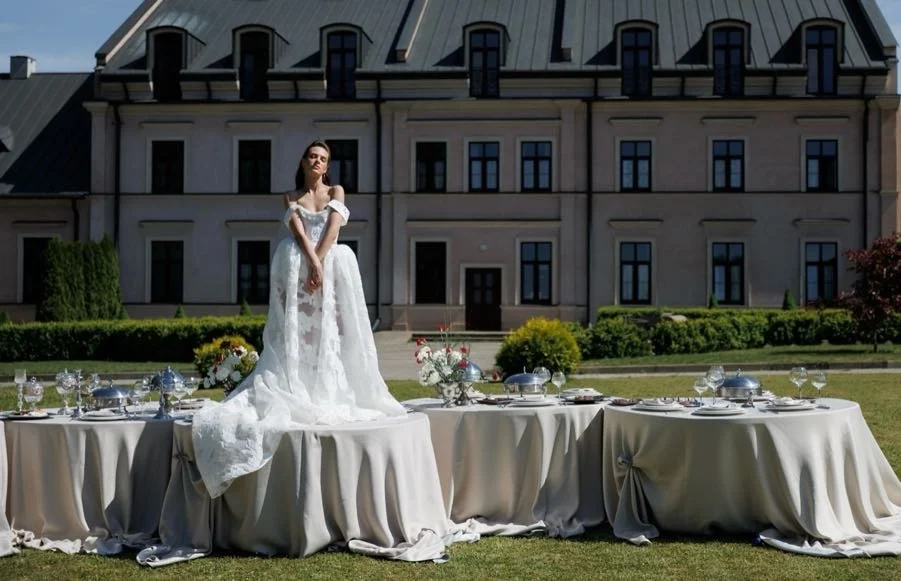 A woman in a white wedding dress standing on a table set for a formal outdoor event in front of a large building.