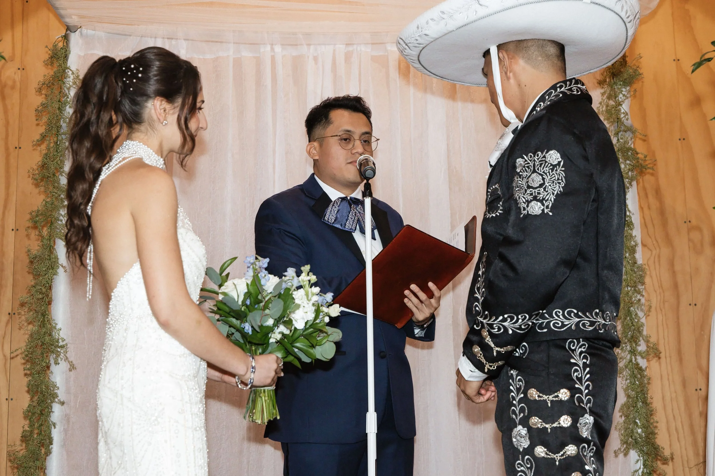 A wedding ceremony with a bride in a white dress holding a bouquet and a groom in a black embroidered outfit with a large sombrero, standing before an officiant who is reading from a document.