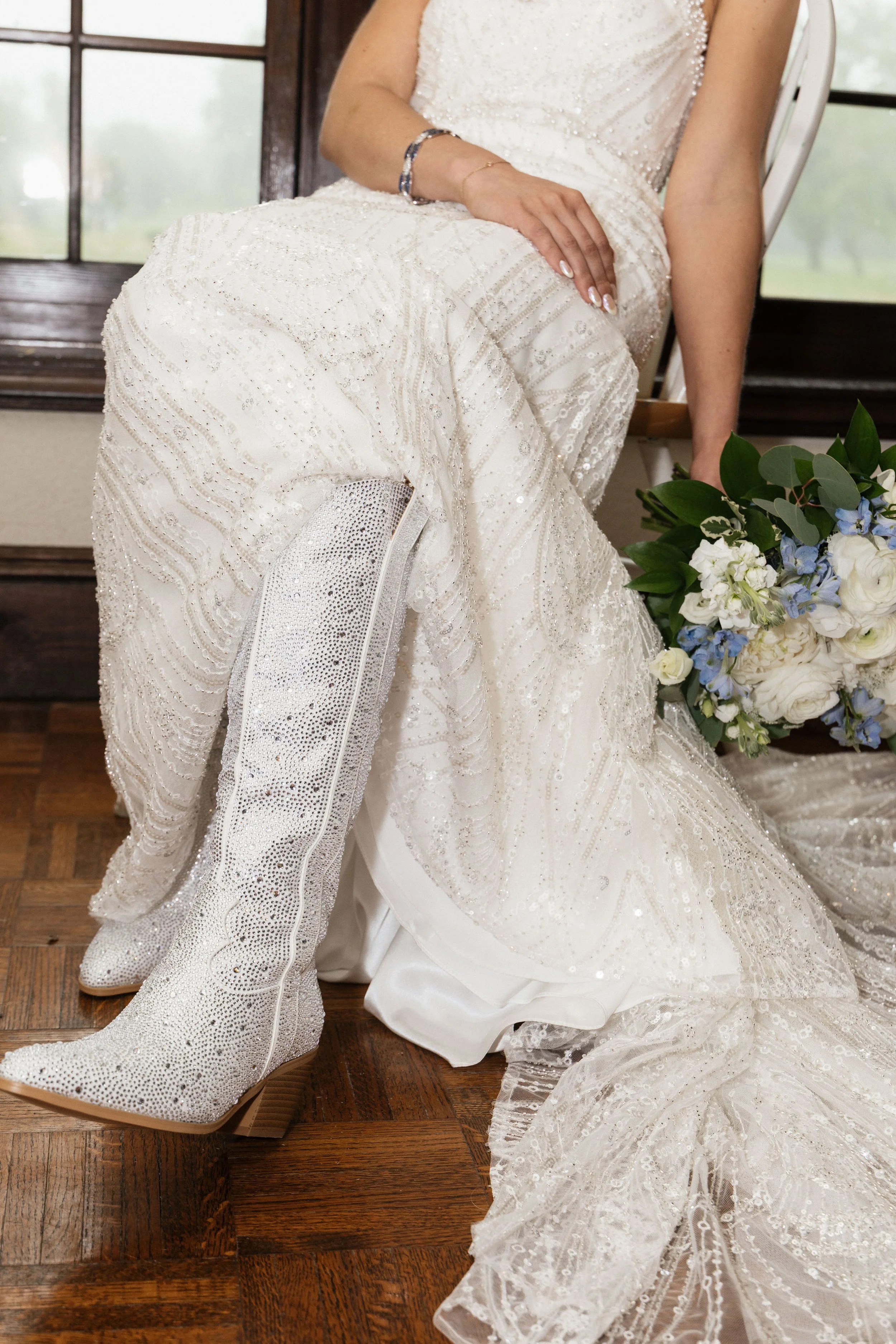 Woman in an elegant white wedding dress and matching high-heeled boots sitting on a chair near a window with natural light, holding a bouquet of blue and white flowers.