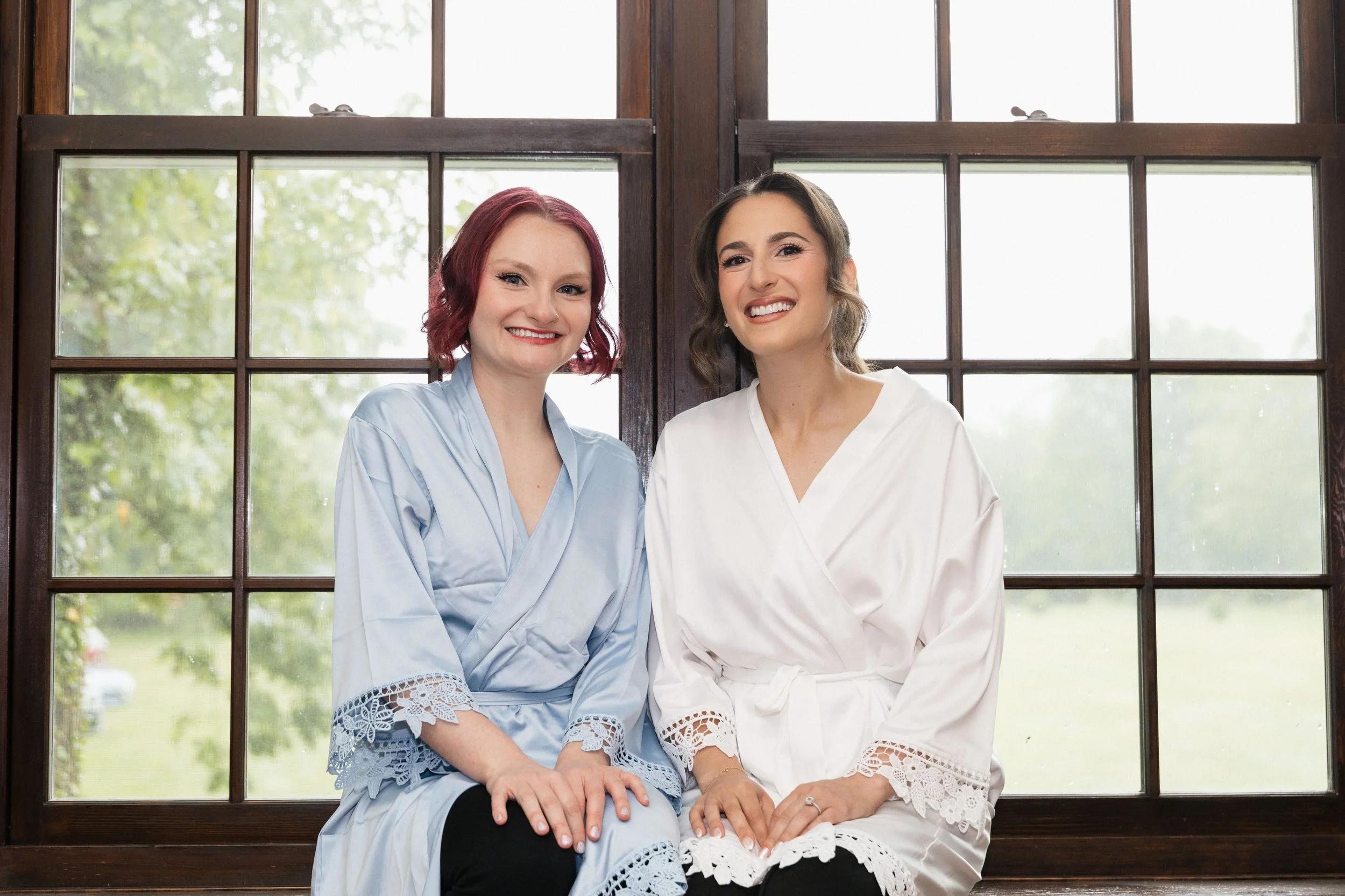 Two women sitting on a windowsill, smiling. One has light skin and red hair, wearing a light blue dress with lace detailing. The other has light skin and dark brown hair with highlights, wearing a white dress with lace accents.