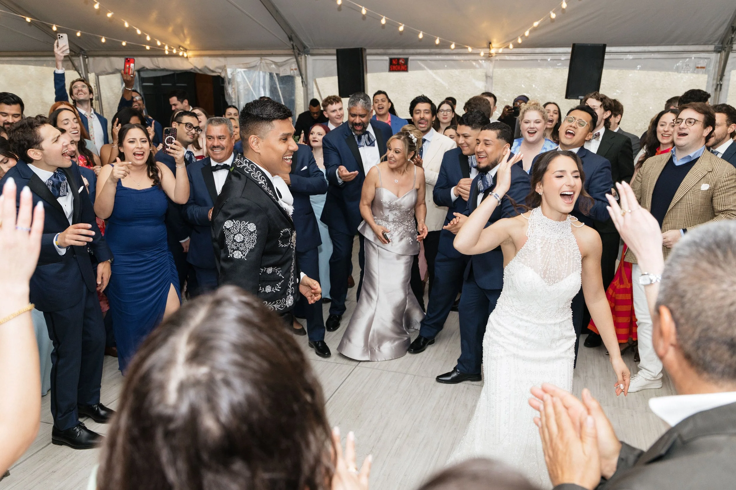 A large group of people dressed in formal attire are dancing and celebrating at an indoor wedding reception with string lights overhead.