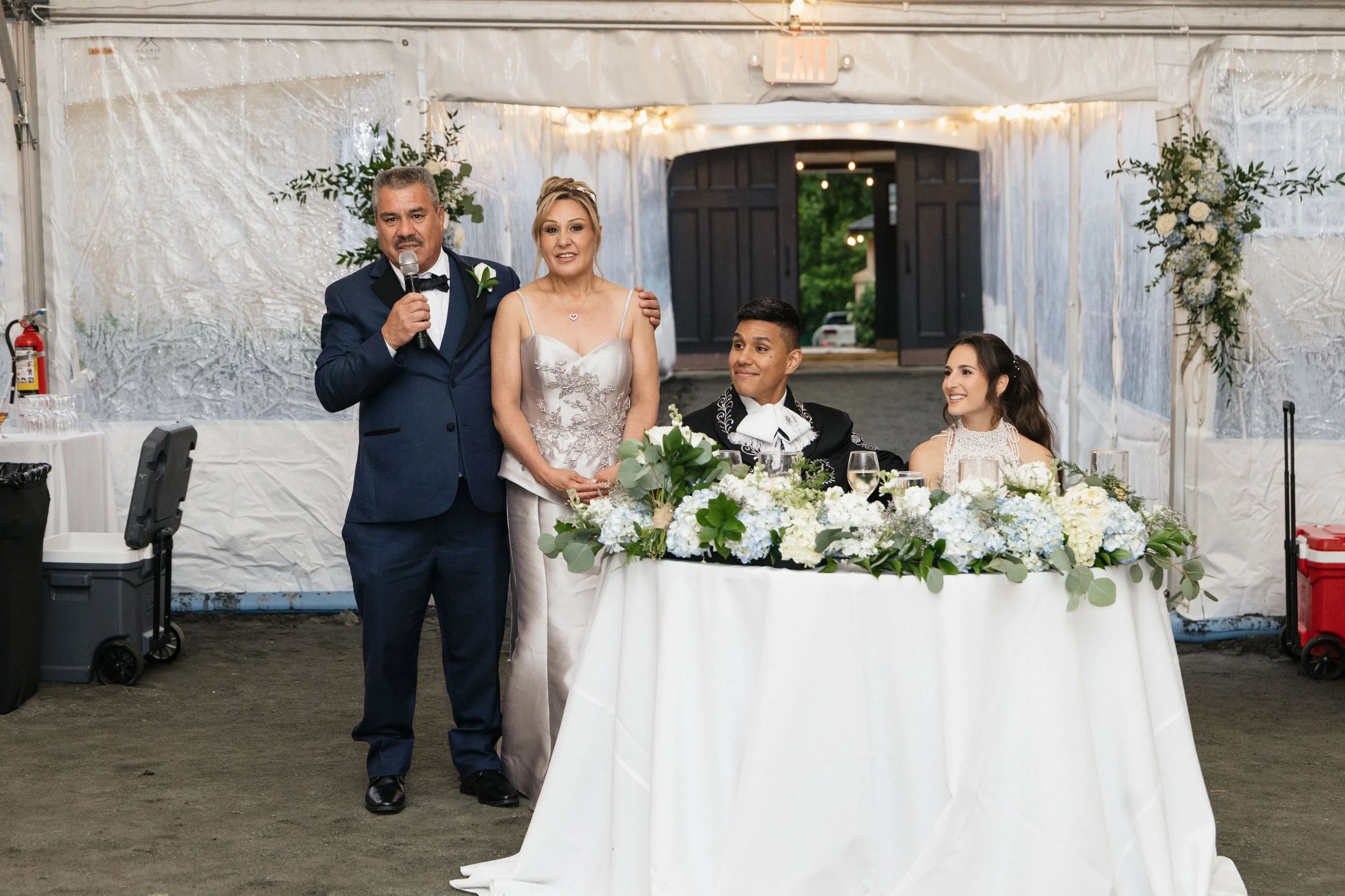 A group of four people at a wedding reception, with a man in a tuxedo speaking into a microphone, a woman in a white dress standing beside him, a groom in a black suit sitting at a decorated table, and a woman with long dark hair sitting next to him,