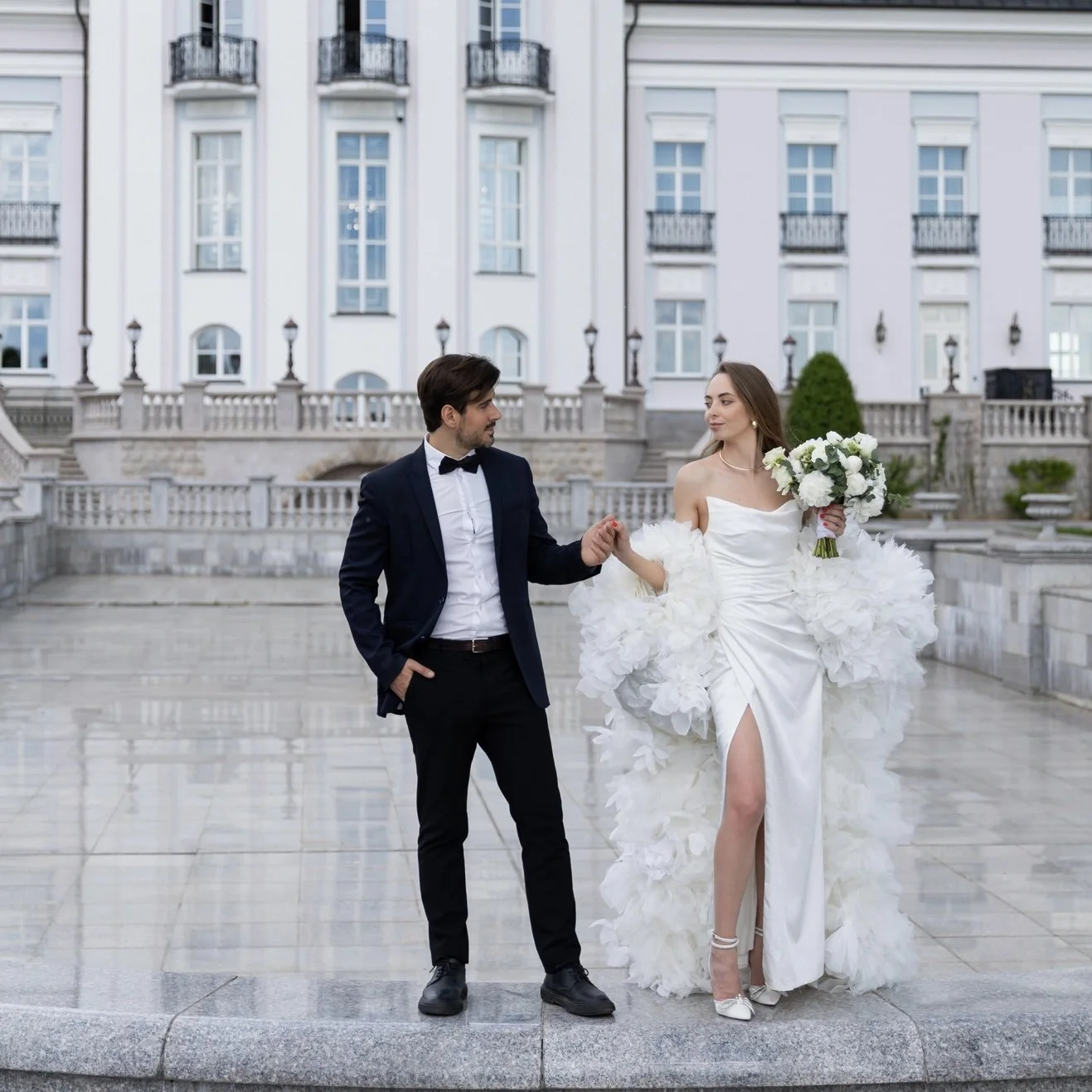 A bride and groom walking hand in hand outdoors in front of a large elegant building, wedding photo, woman in a white wedding gown holding a bouquet, man in a black tuxedo, moment of connection, romantic atmosphere.