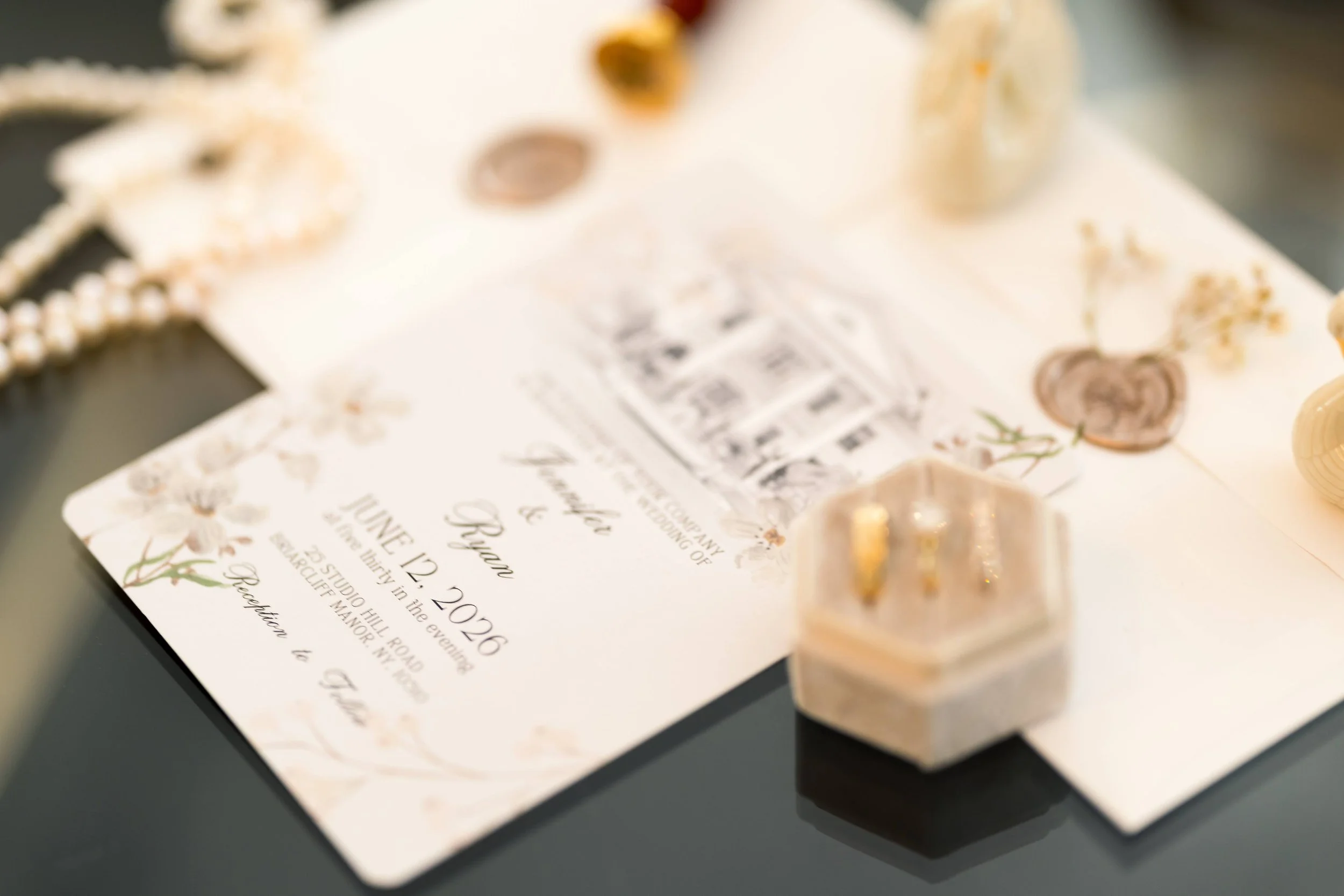 Close-up of a wedding invitation card with decorative floral illustrations, along with gold jewelry pieces and a pearl necklace placed on top.