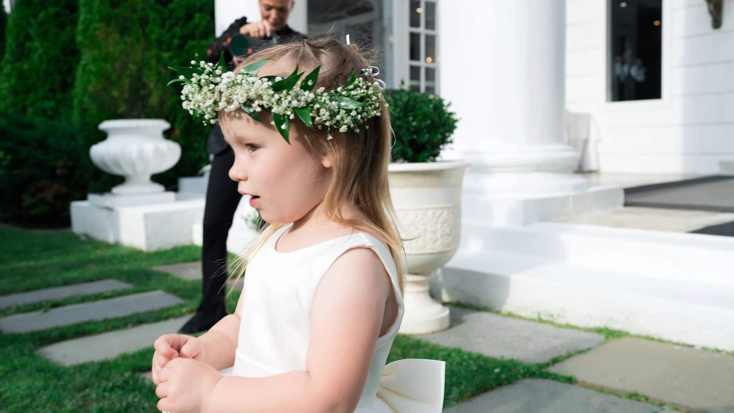 A young girl with long blonde hair wearing a white dress and a flower crown made of small white flowers and green leaves, standing outdoors in front of a white building with stairs and a large planter, with another person in the background.