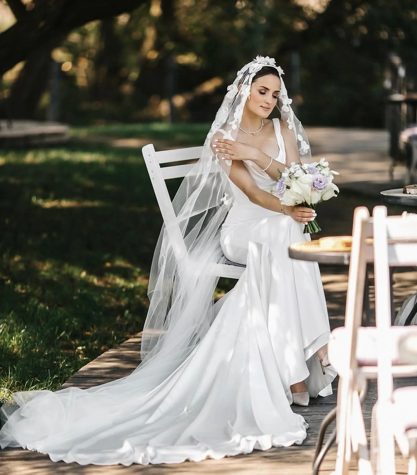 A bride sitting on a white chair outdoors, wearing a white wedding dress and veil, holding a bouquet of white and purple flowers, with her hand on her shoulder and her eyes closed.