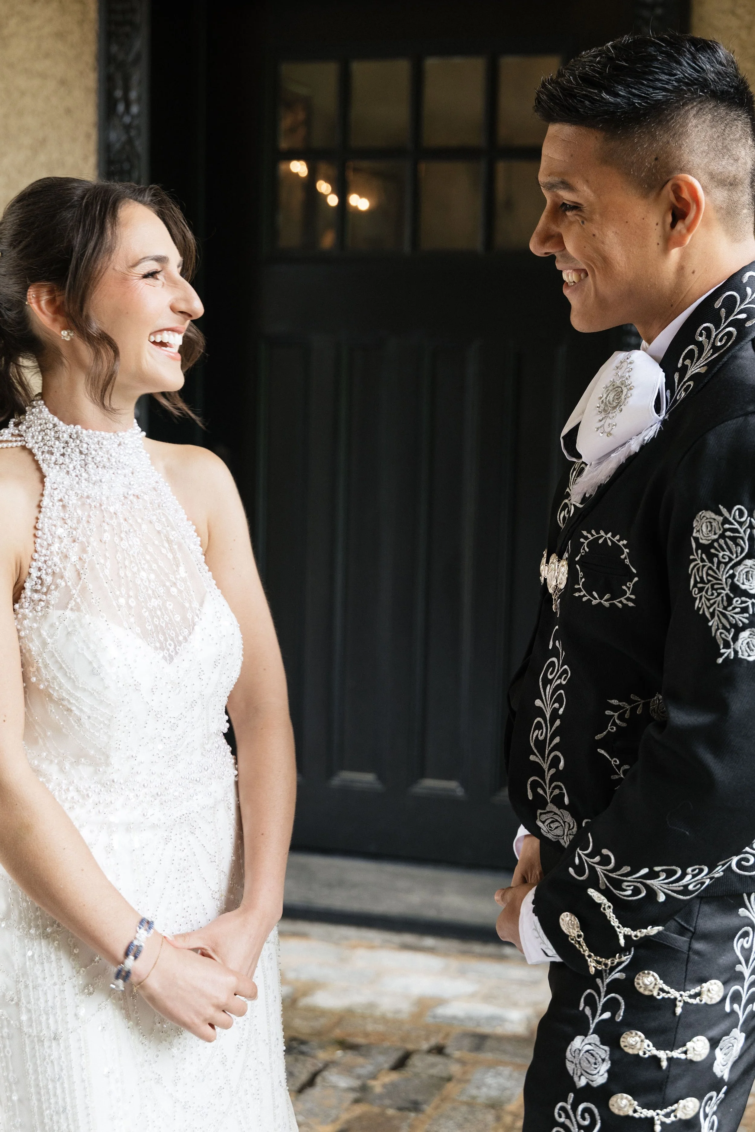 A bride and groom smiling at each other during their wedding ceremony, standing outdoors in front of a black door.