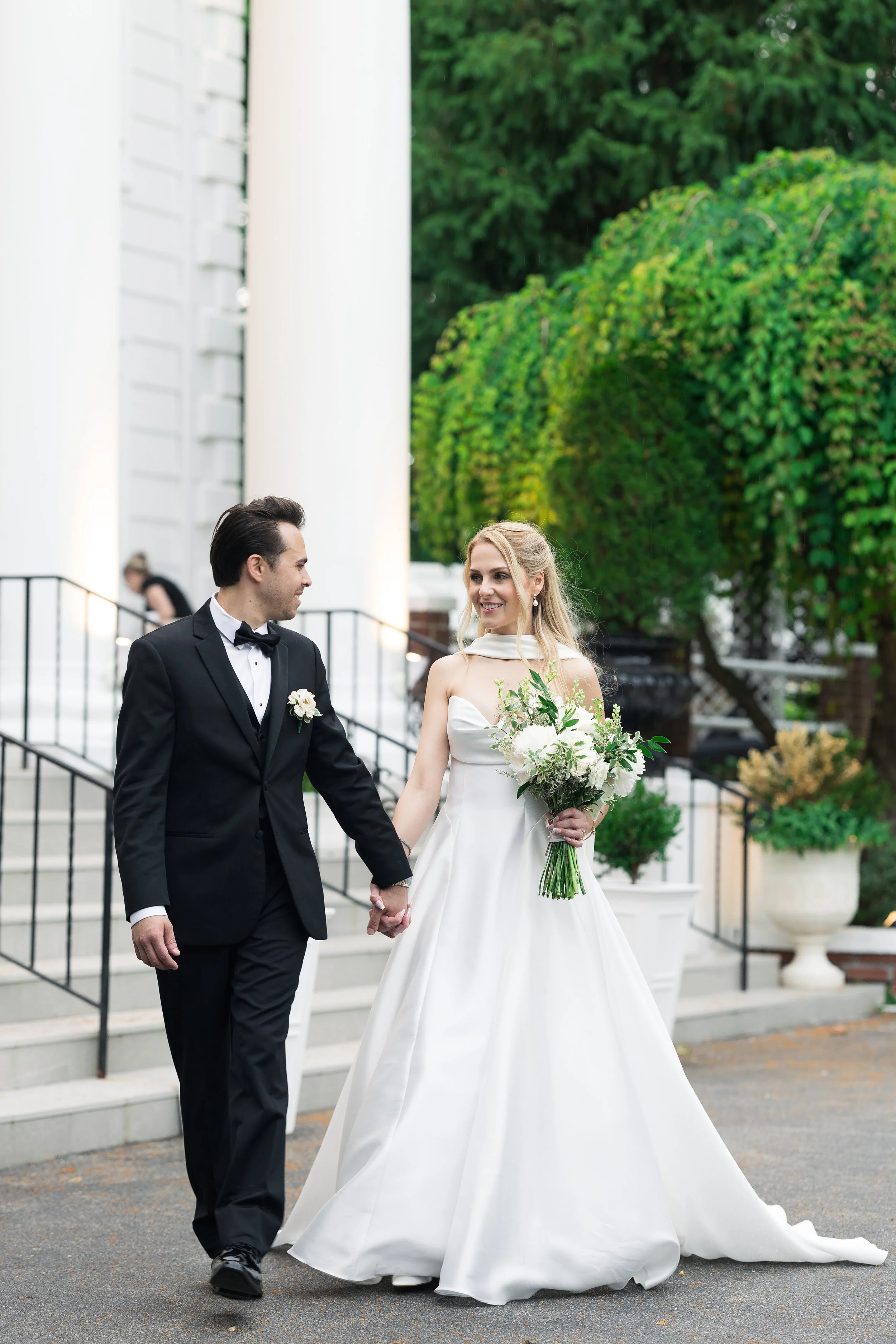 A bride and groom holding hands and walking outside on stairs, smiling and looking at each other. The groom is wearing a black tuxedo with a bow tie, and the bride is in a white wedding gown holding a bouquet of white and green flowers. There is gree