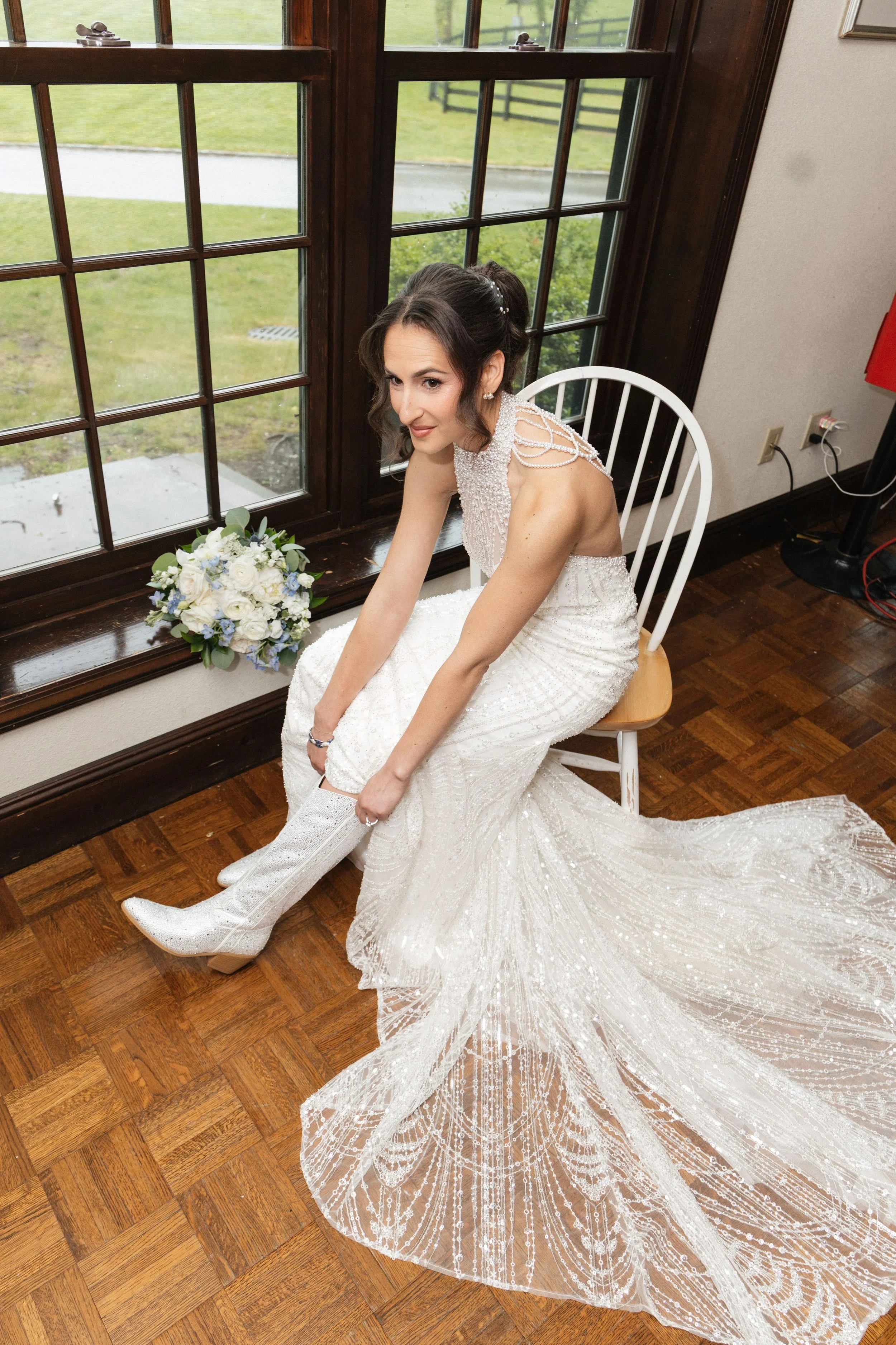 A bride in a white dress sitting on a chair by a large window, with a bouquet of white and blue flowers beside her, adjusting her white high heel shoe, with a wedding dress train spread out on the wooden floor.