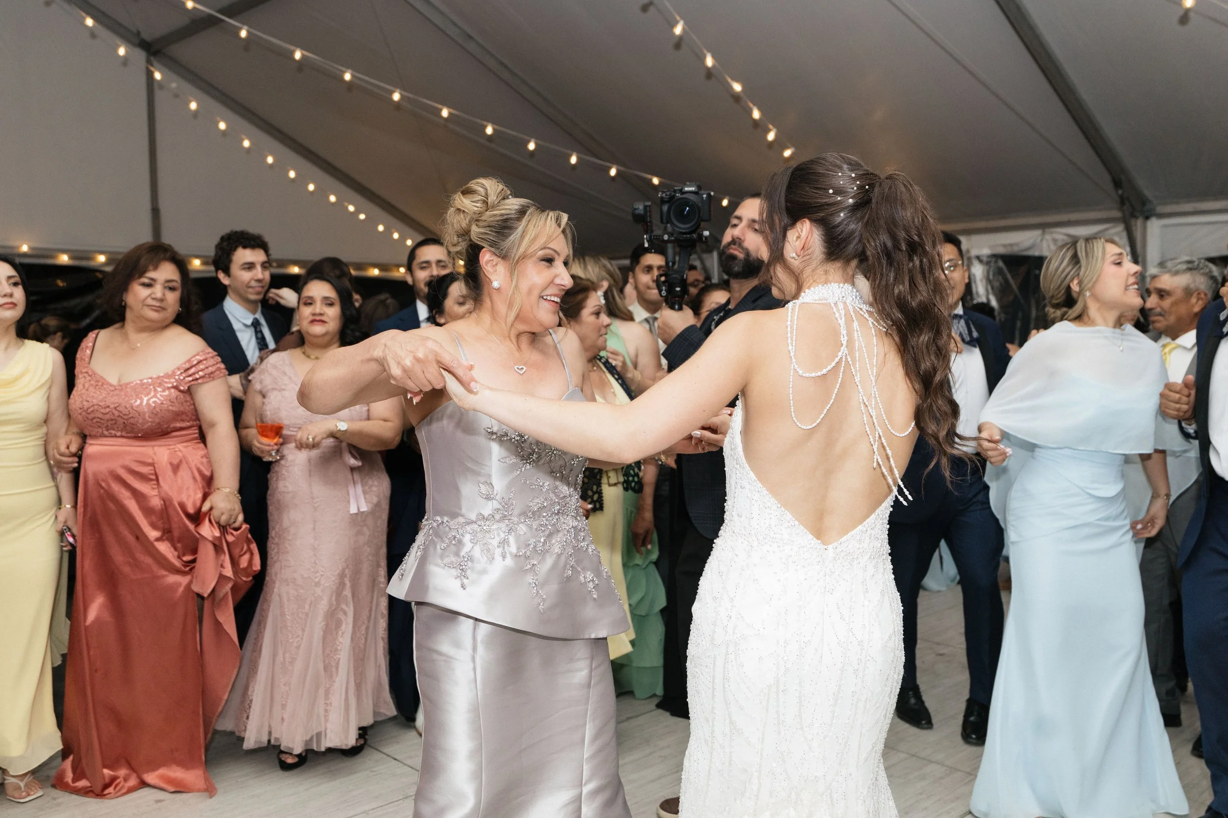 People dancing at a wedding reception inside a decorated tent, with string lights overhead. The focus is on two women, one in a light gray dress and the other in a white dress, holding hands and smiling at each other.