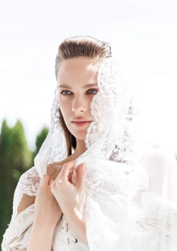 A young woman with blue eyes and slicked-back hair, wearing a white lace shawl over her head, standing outdoors with greenery in the background.