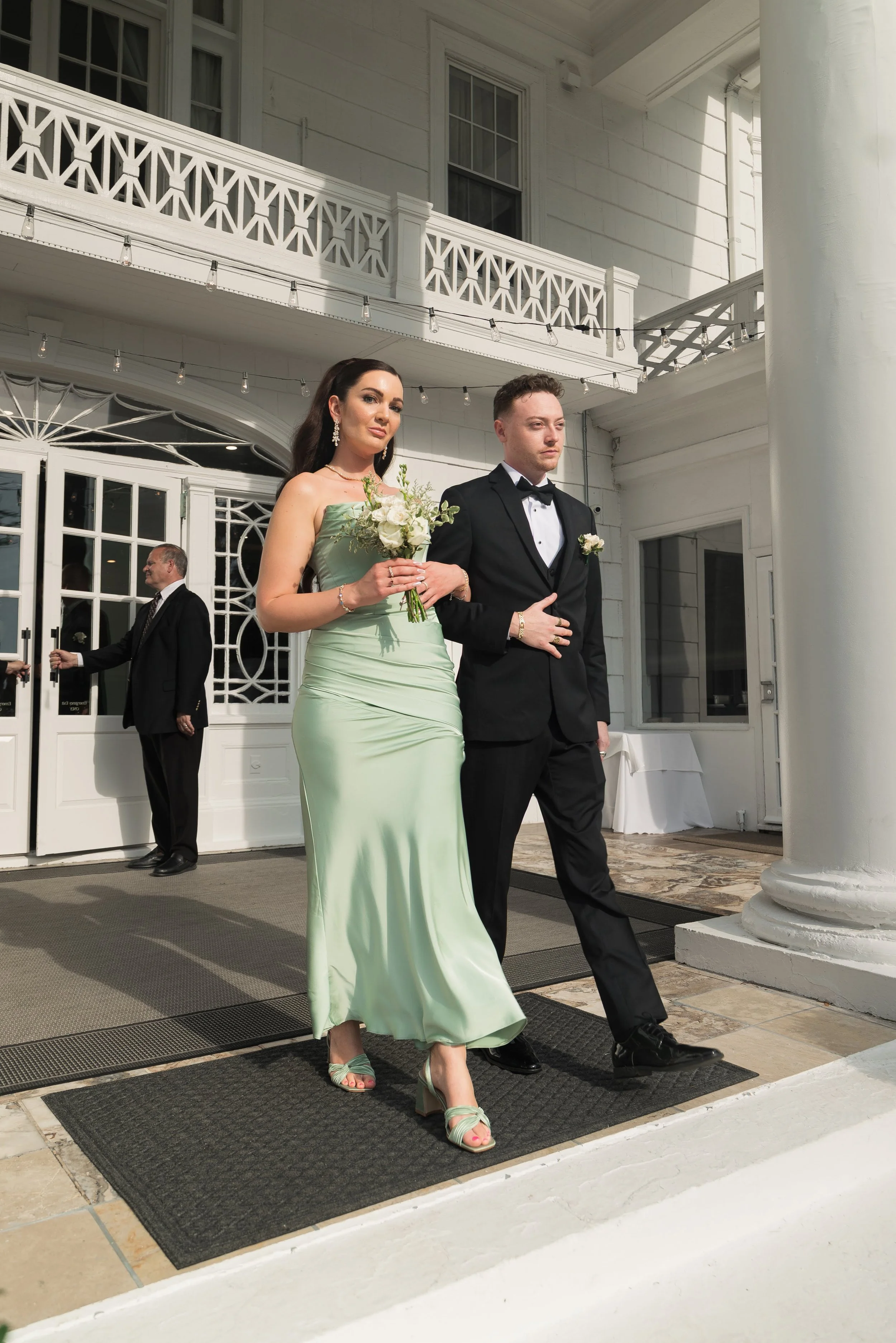 A bride and groom walking out of a building with white walls and columns, with the bride holding a bouquet of flowers. The bride is wearing a light green strapless dress and matching heels, and the groom is in a black tuxedo with a bow tie. There are