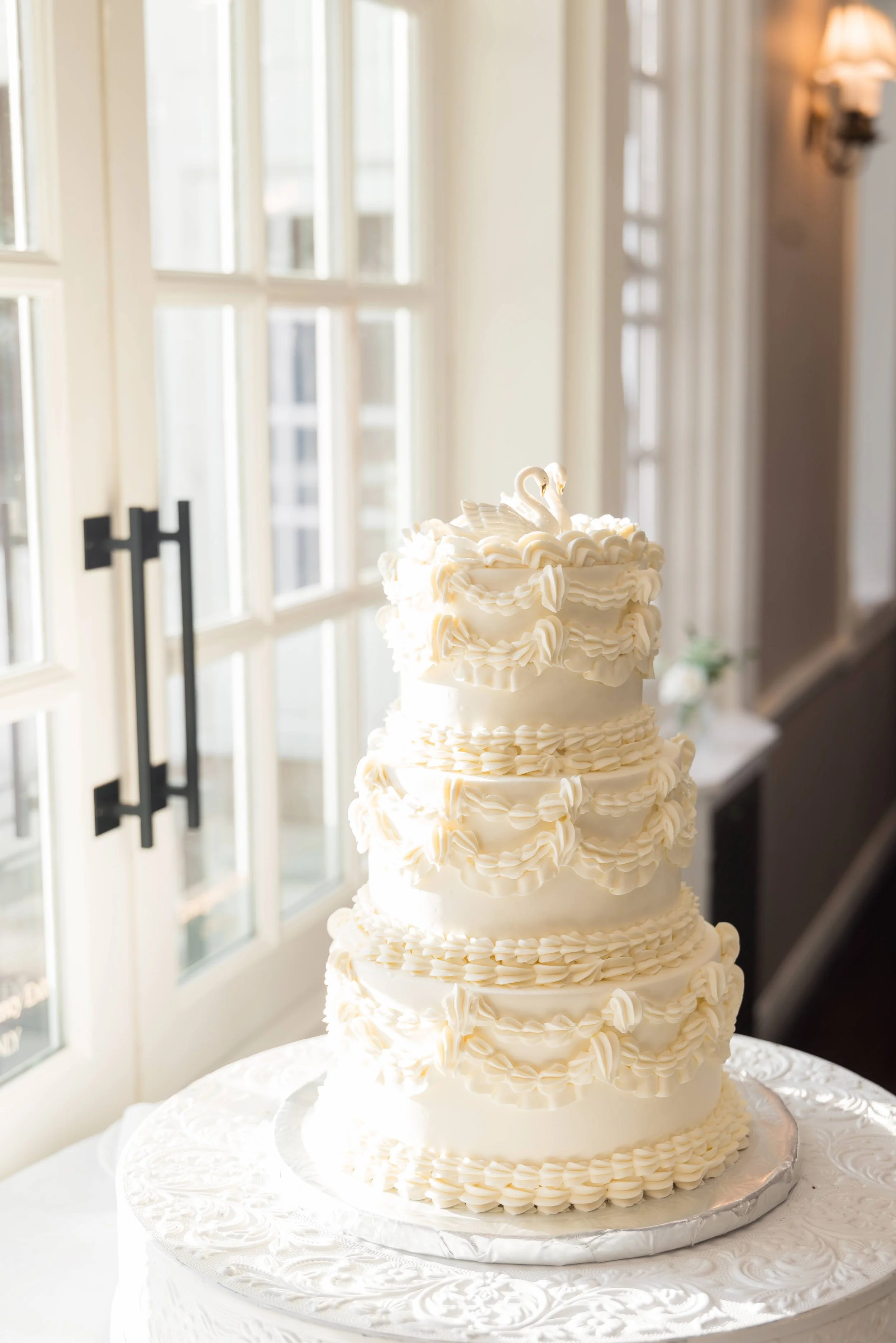 A tall white wedding cake with multiple layers, decorated with piped buttercream and two swan toppers, placed on a white ornate tablecloth in front of a window with daylight streaming in.