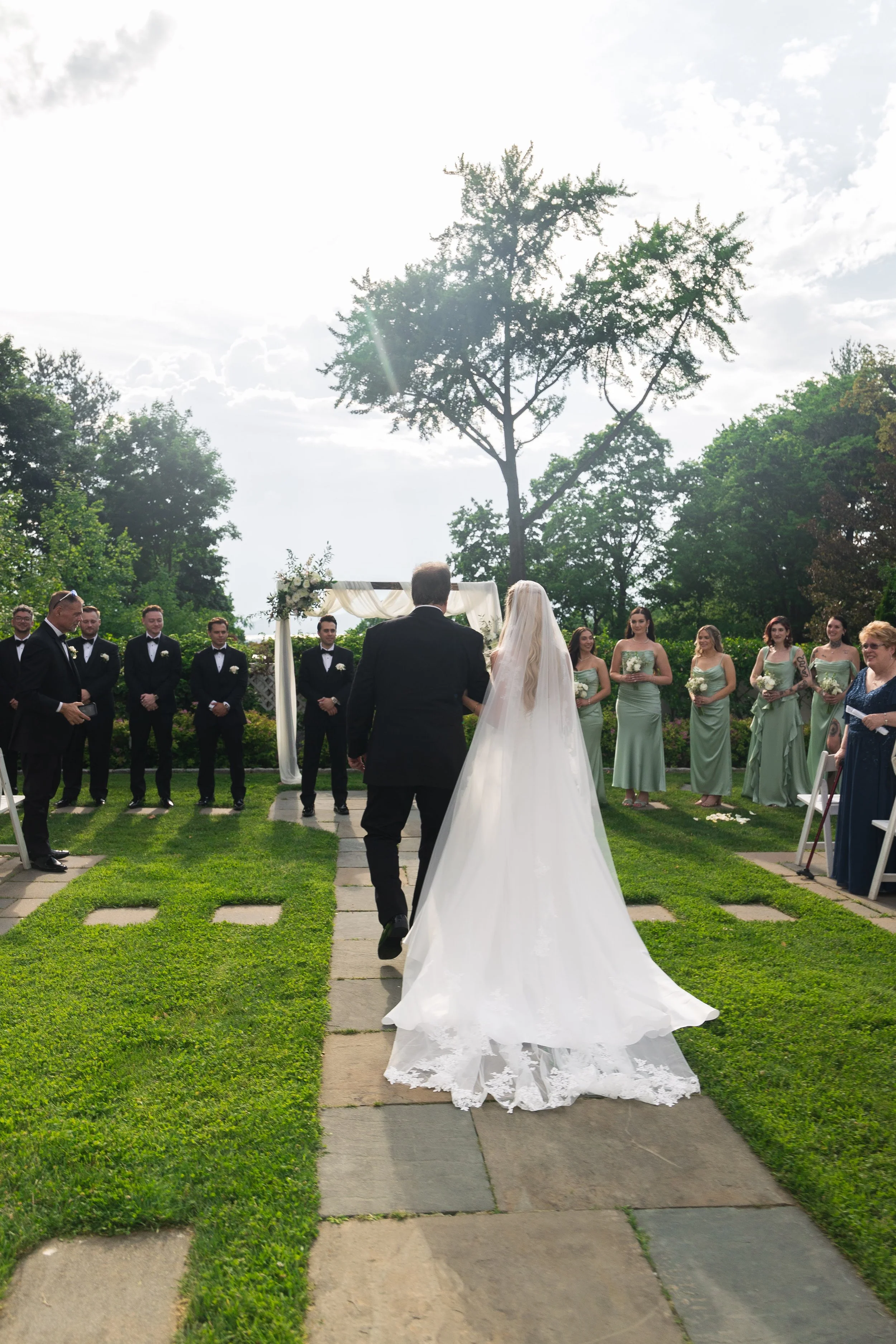 A bride in a white wedding gown walking down an outdoor aisle towards a groom, with bridesmaids in green dresses and groomsmen in tuxedos standing on either side, under a floral arch in a garden setting with trees and cloudy sky.