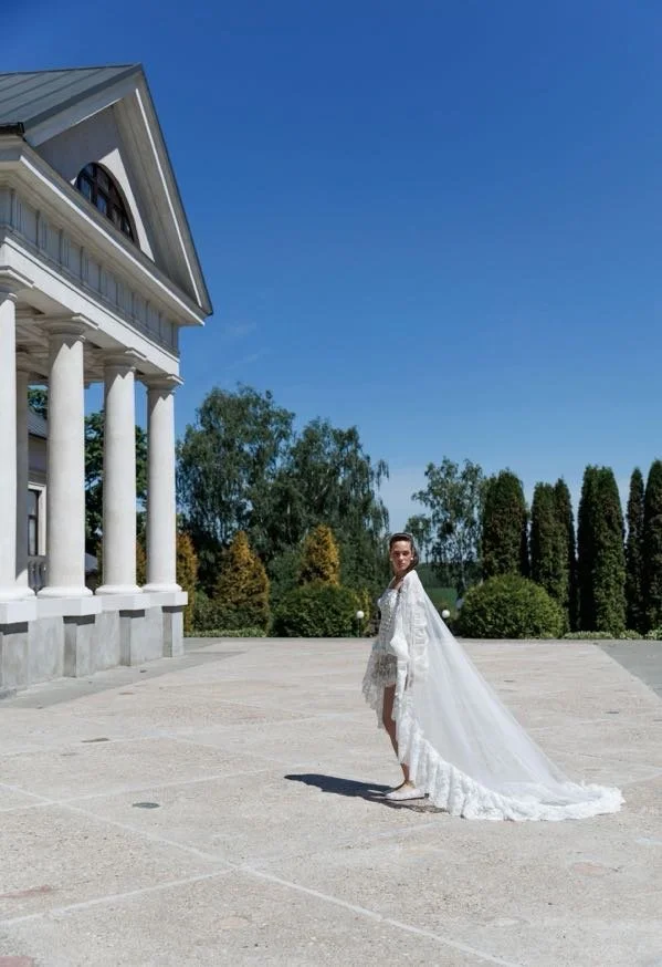 A woman in a white wedding dress with a long train standing outside on a sunny day, near a building with classical columns, with trees and a clear blue sky in the background.
