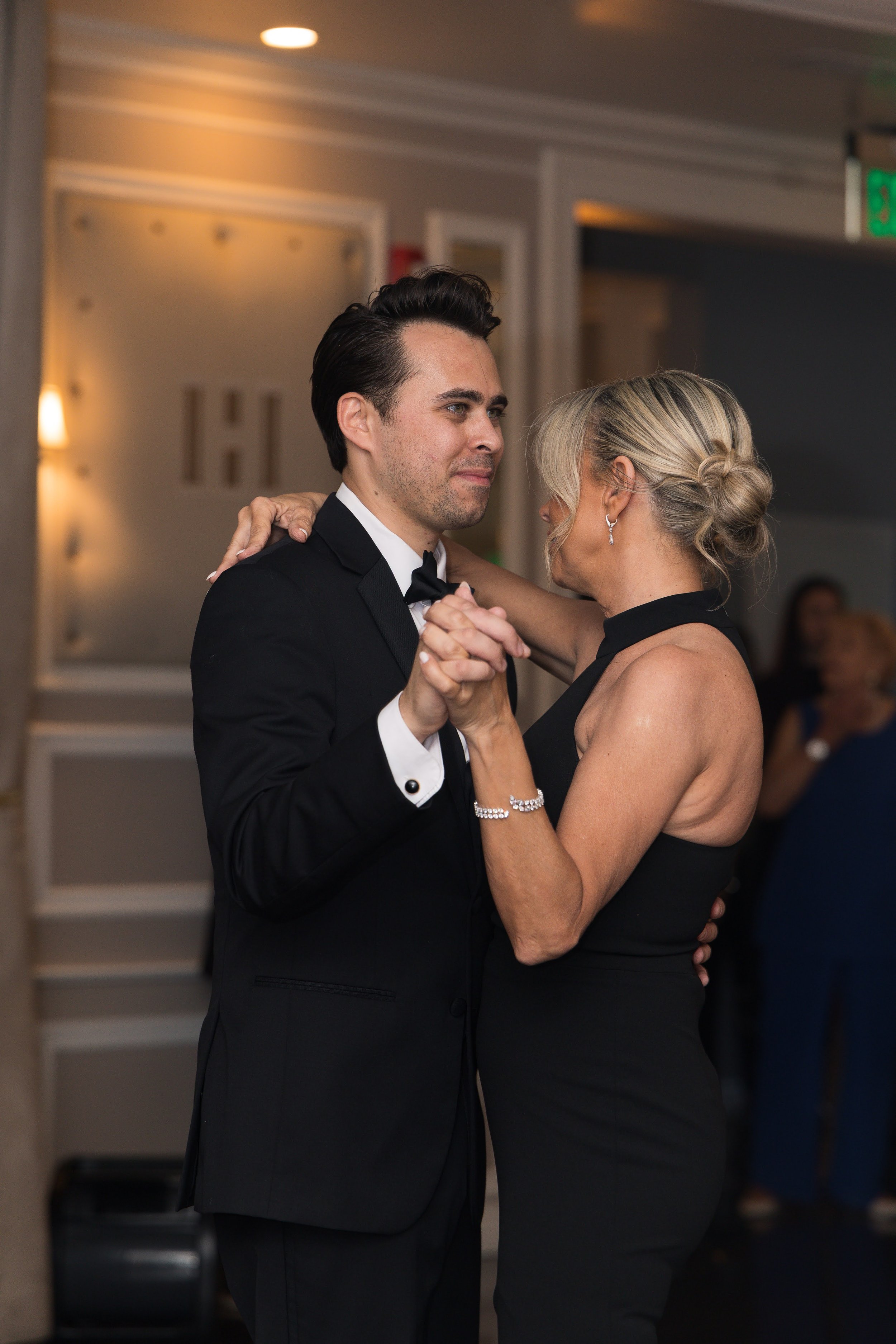 A man and a woman dancing closely at a formal event. The man is in a black tuxedo and the woman is in a black dress. They are holding hands and looking at each other.