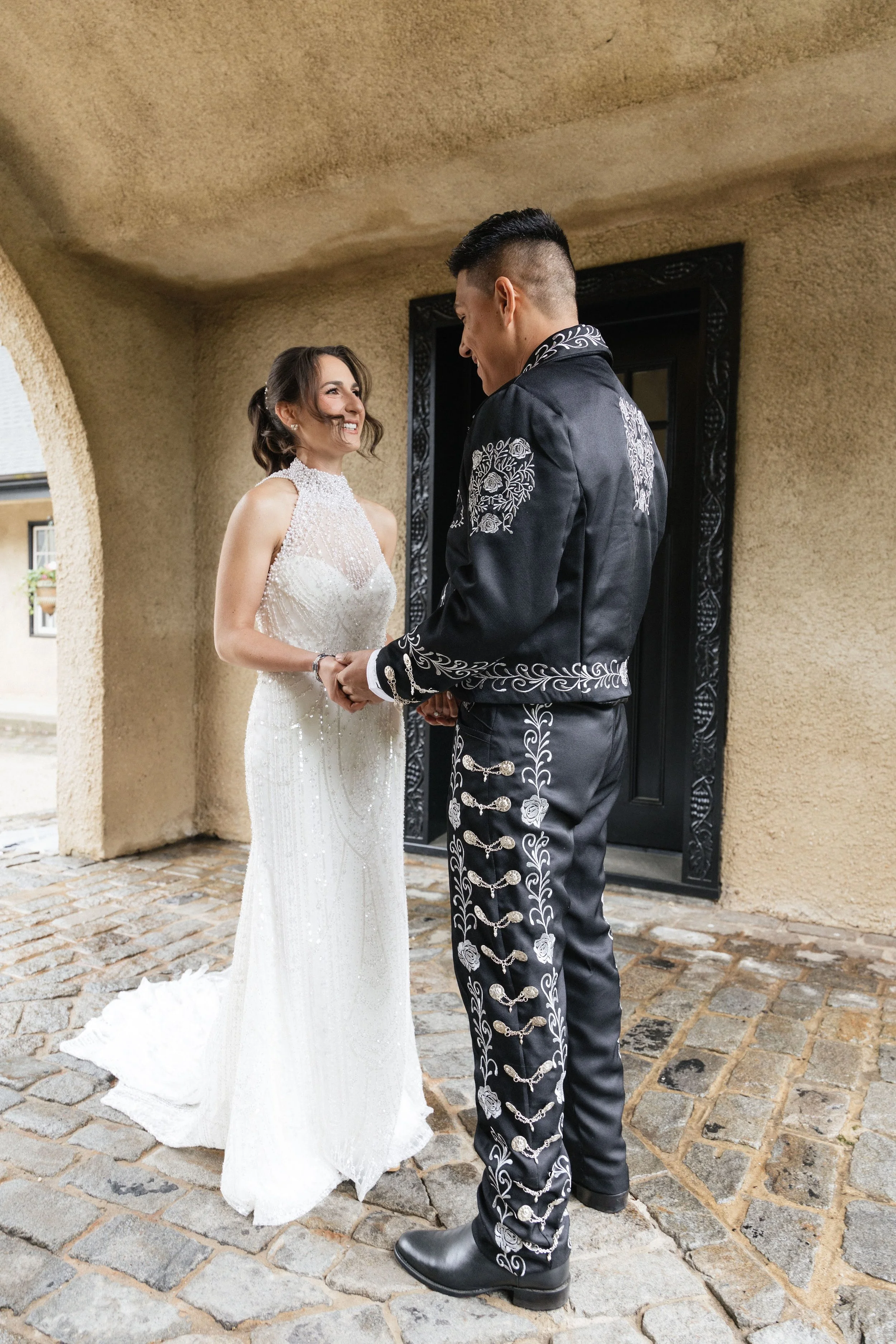 A bride and groom holding hands and smiling at each other outside a building with a rustic stone-paved ground and a textured beige wall.