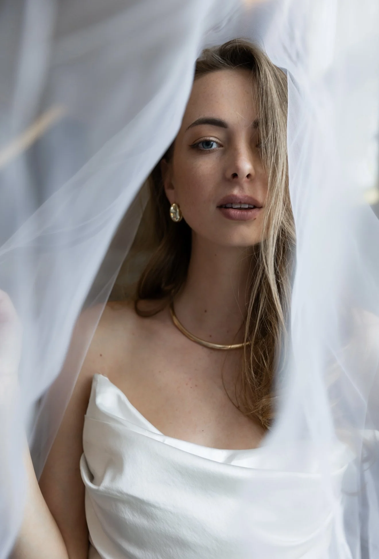 A young woman with blonde hair and blue eyes is partially covered by semi-transparent white fabric, revealing her face and upper chest, dressed in white satin-like clothing, with a gold necklace and earrings.