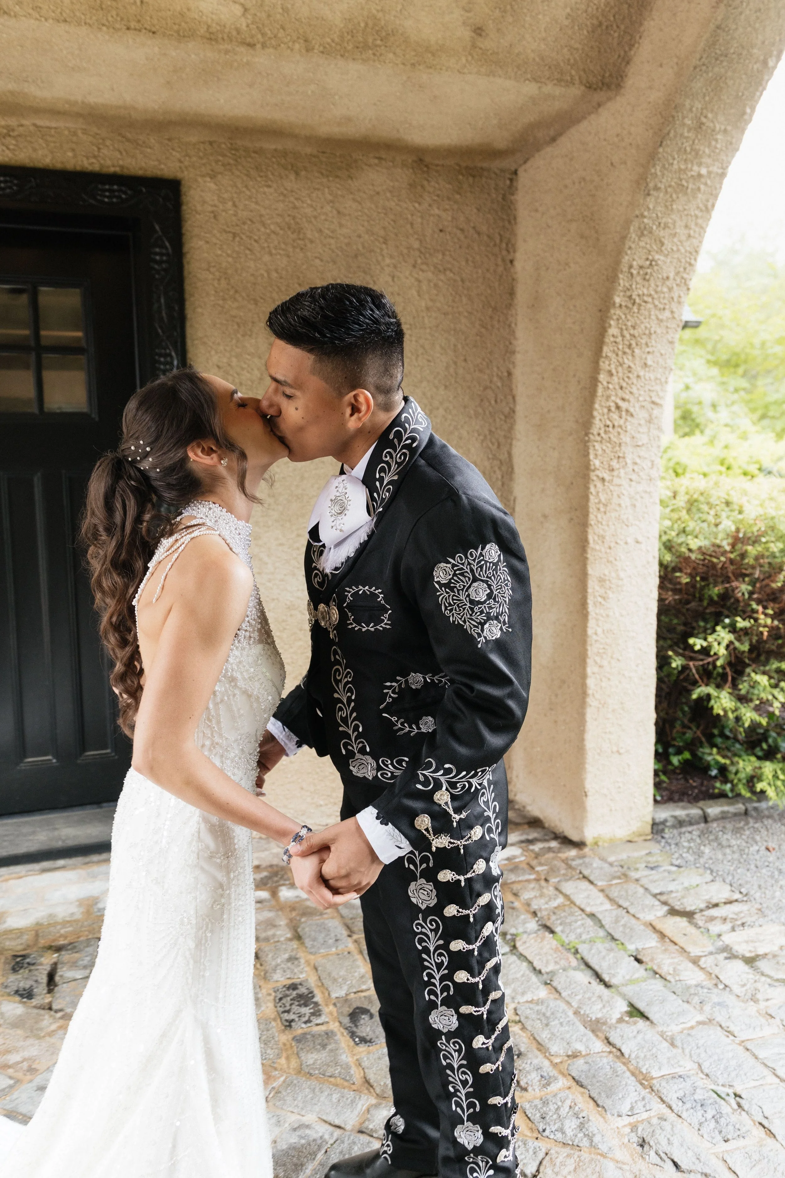 A couple in wedding attire sharing a kiss while holding hands outside a building with a cobblestone path.