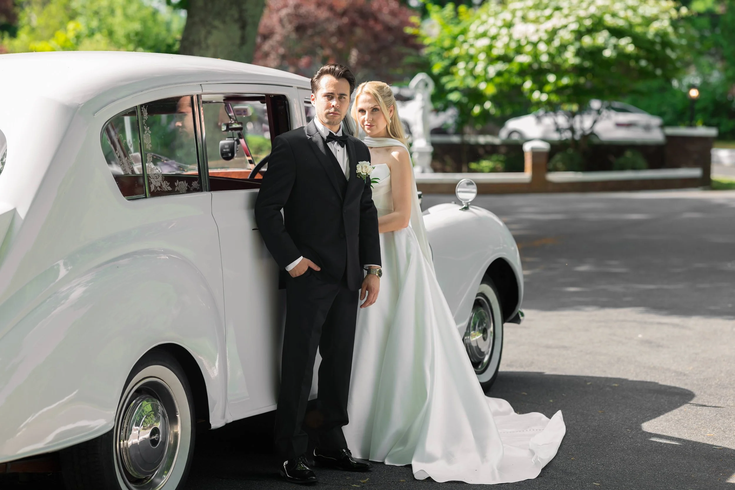 A bride and groom stand next to a white vintage car during a wedding, with trees and a fence in the background.