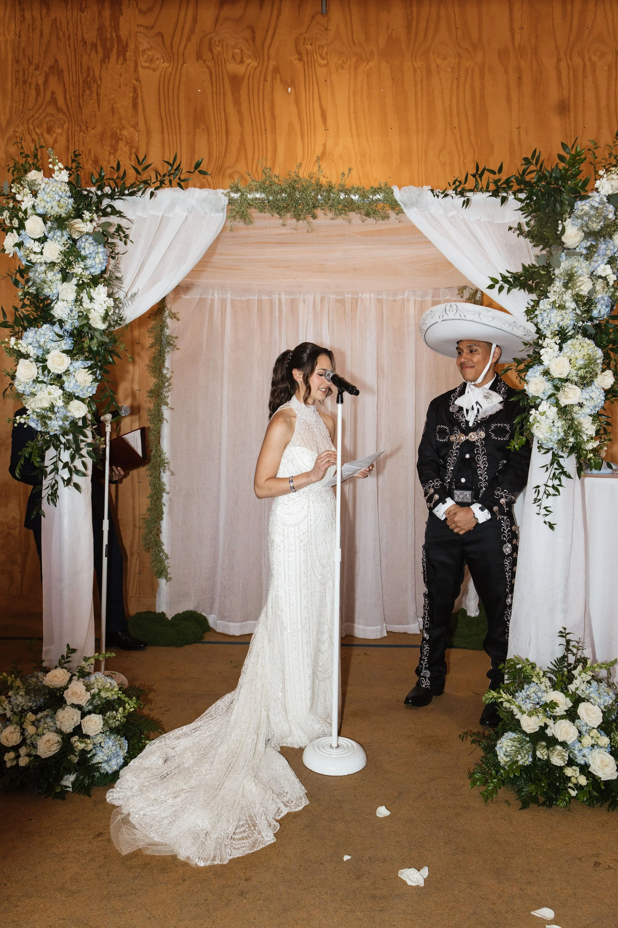 A bride and groom standing under a floral wedding arch with white curtains, during a wedding ceremony. The bride wears a white lace dress, holding a paper, and the groom wears traditional Mexican attire with a large sombrero. The setting has floral a