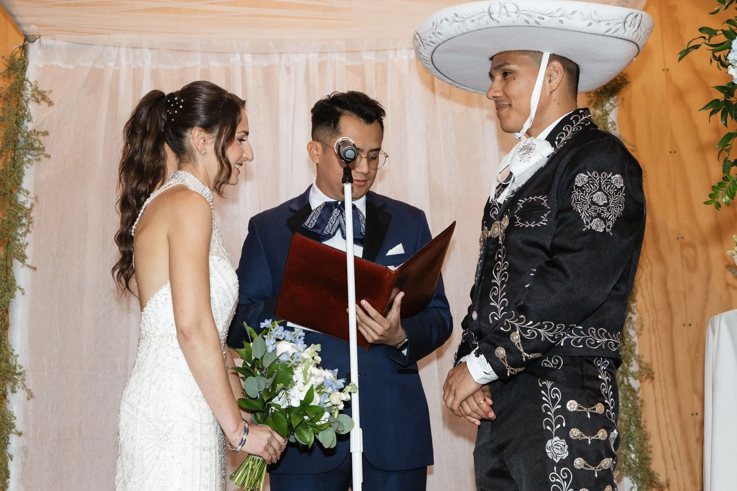 A couple getting married, holding hands and facing each other in front of an officiant, with a decorated backdrop and greenery, during a wedding ceremony.