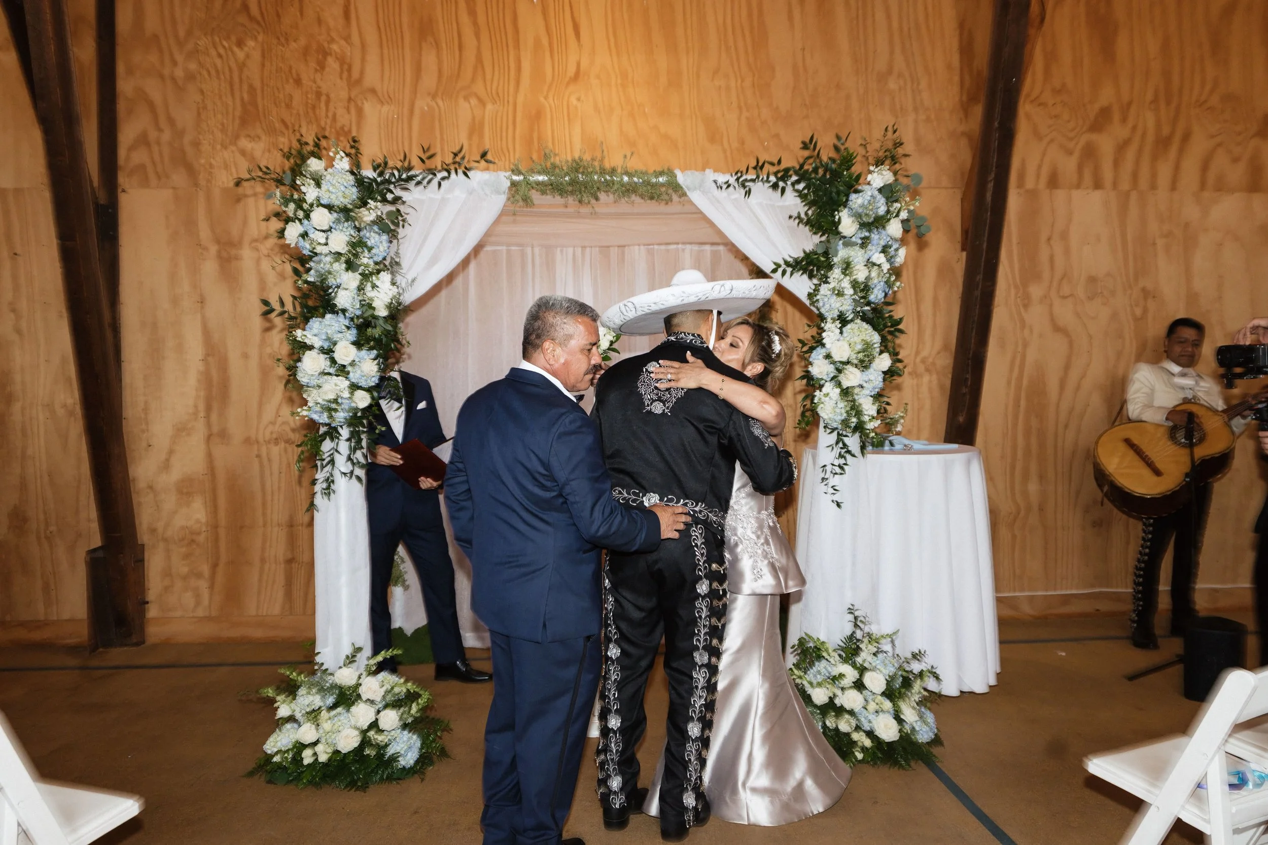 A couple is sharing a kiss during a wedding ceremony on a decorated wooden arch filled with white and blue flowers, with two men standing nearby, one playing guitar and another holding a book or folder.