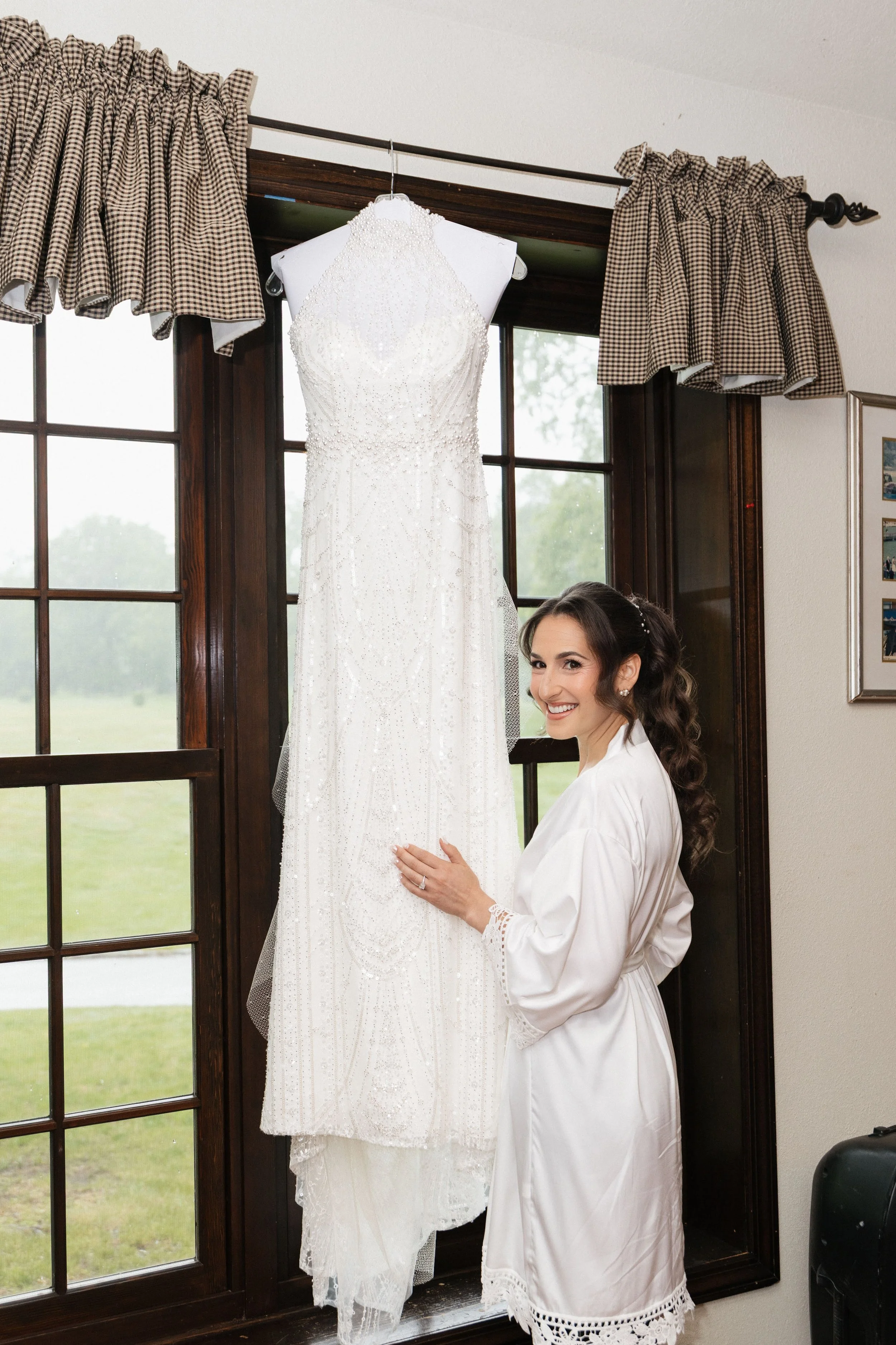 A woman in a satin robe is smiling and touching a white wedding dress hanging in front of a large window with brown wooden frames and checkered curtains.