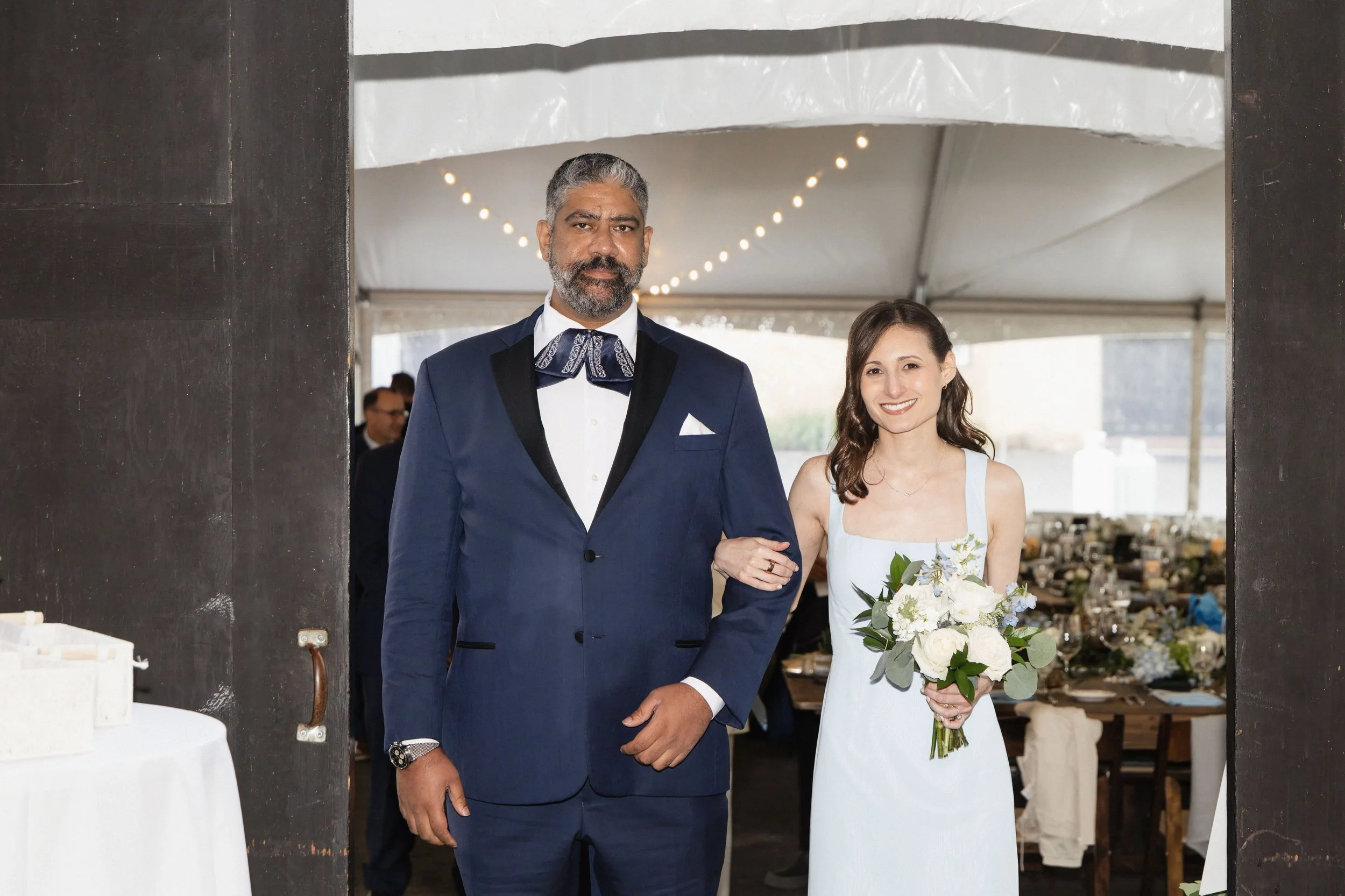 A bride and groom standing together at their wedding reception, arms linked. The groom wears a navy blue tuxedo with a white shirt and a patterned bow tie, while the bride wears a white wedding dress and holds a bouquet of white flowers and greenery.
