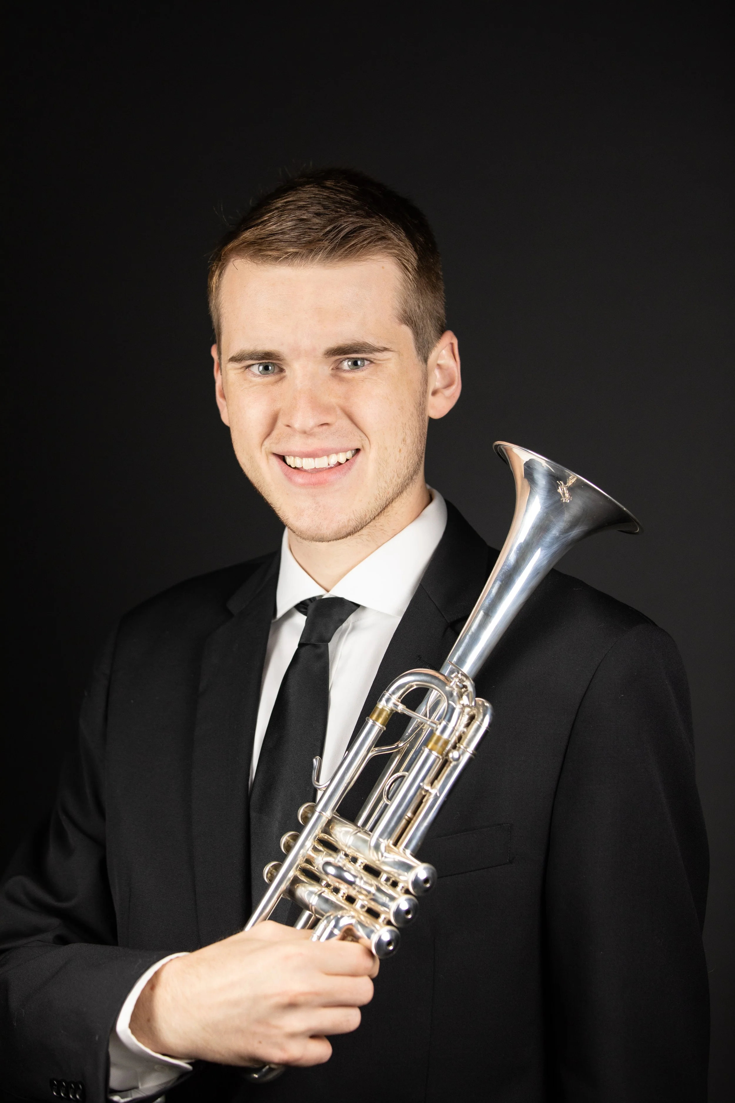 A young man in a black tuxedo holding a silver trumpet, smiling at the camera against a solid black background.