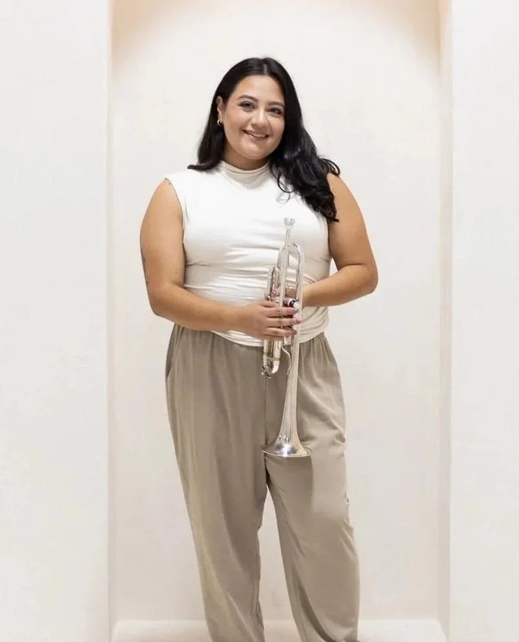 A woman with long black hair, smiling, holding a silver trumpet, dressed in a sleeveless white top and beige pants, standing in front of a plain beige background.