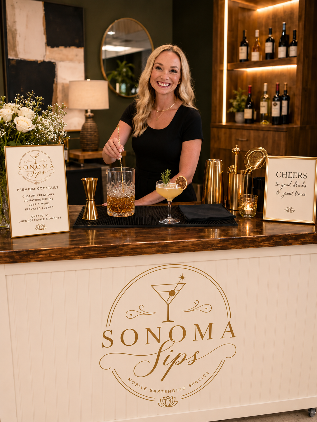 A smiling woman behind a bar counter preparing a cocktail at Sonoma Sips, a mobile bartending service. The bar has signs promoting cocktails and celebrations, with bottles of wine on shelves behind her and decorative lighting.