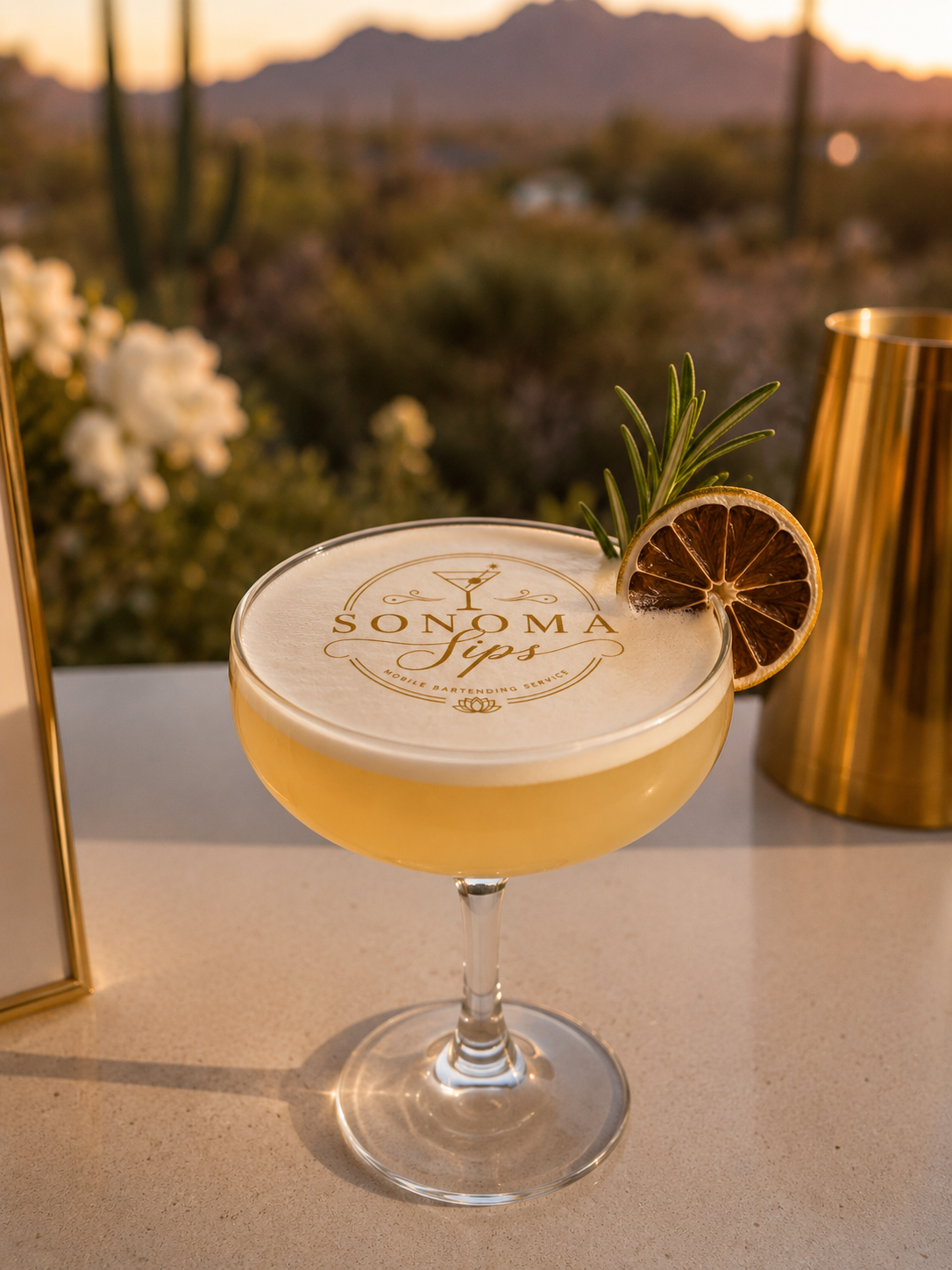A cocktail with a frothy top and garnish of a dried lemon slice and rosemary sprig, on a table at sunset with desert landscape in the background.