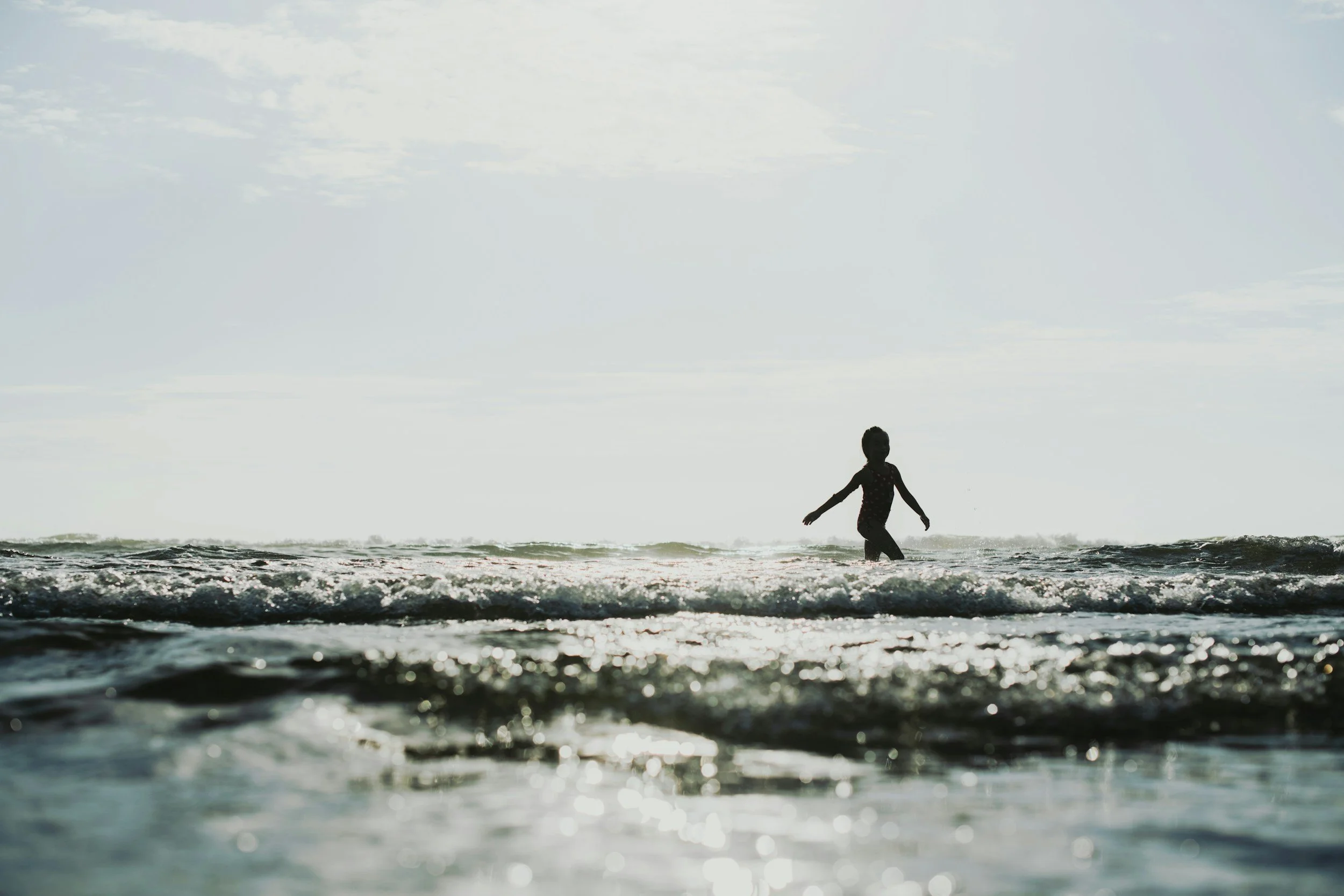 Child playing in the shallow ocean water during sunset or sunrise with the sky and ocean meeting in the background.
