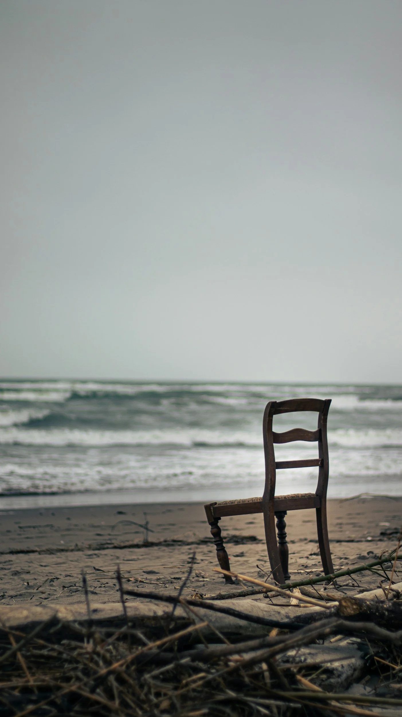 An empty wooden chair on a sandy beach facing the ocean with waves, overcast sky, and some driftwood in the foreground.